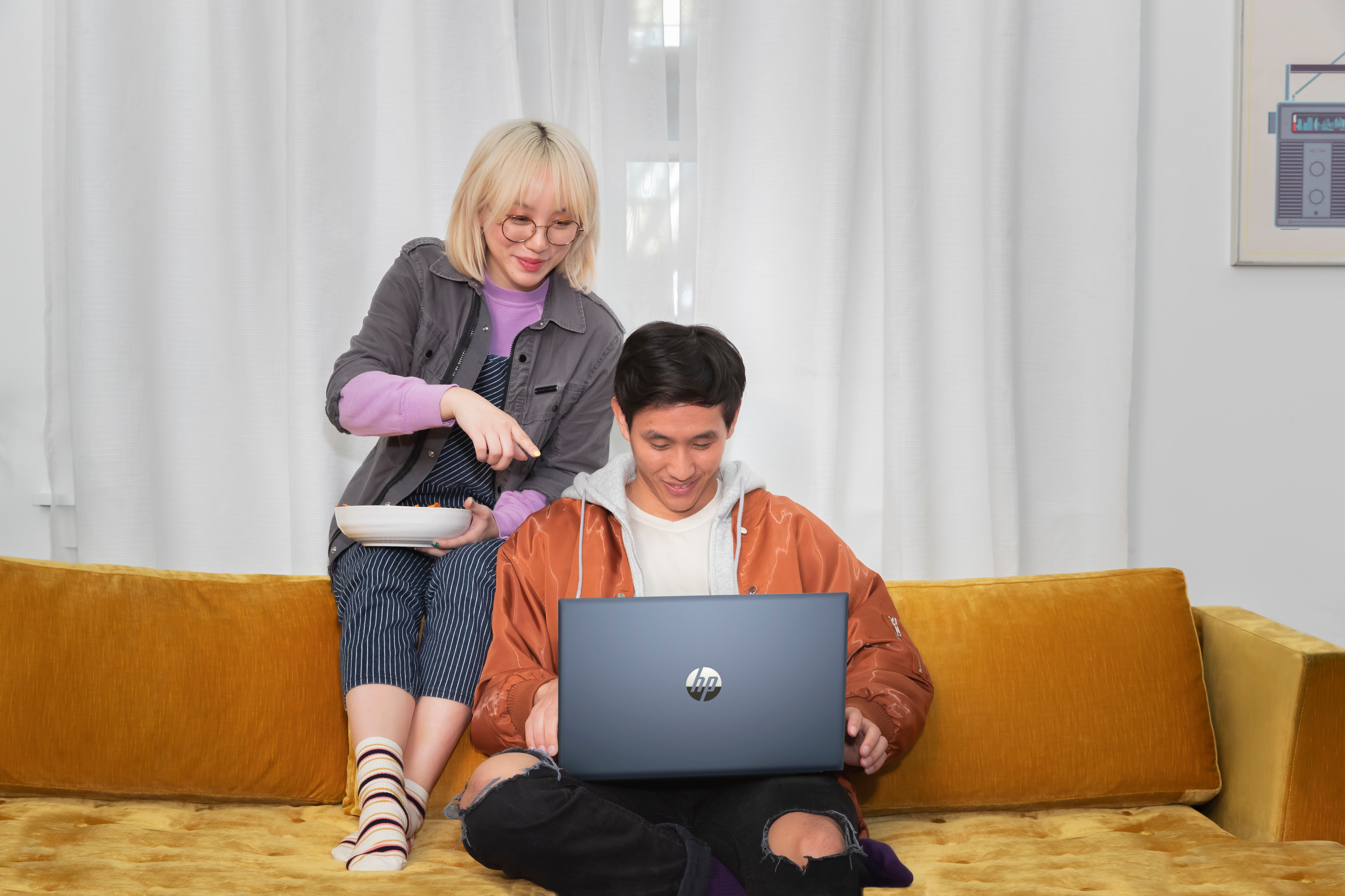 Kobieta trzyma talerz obok mężczyzny używającego laptopa na pomarańczowej sofie. Zasłony w tle.