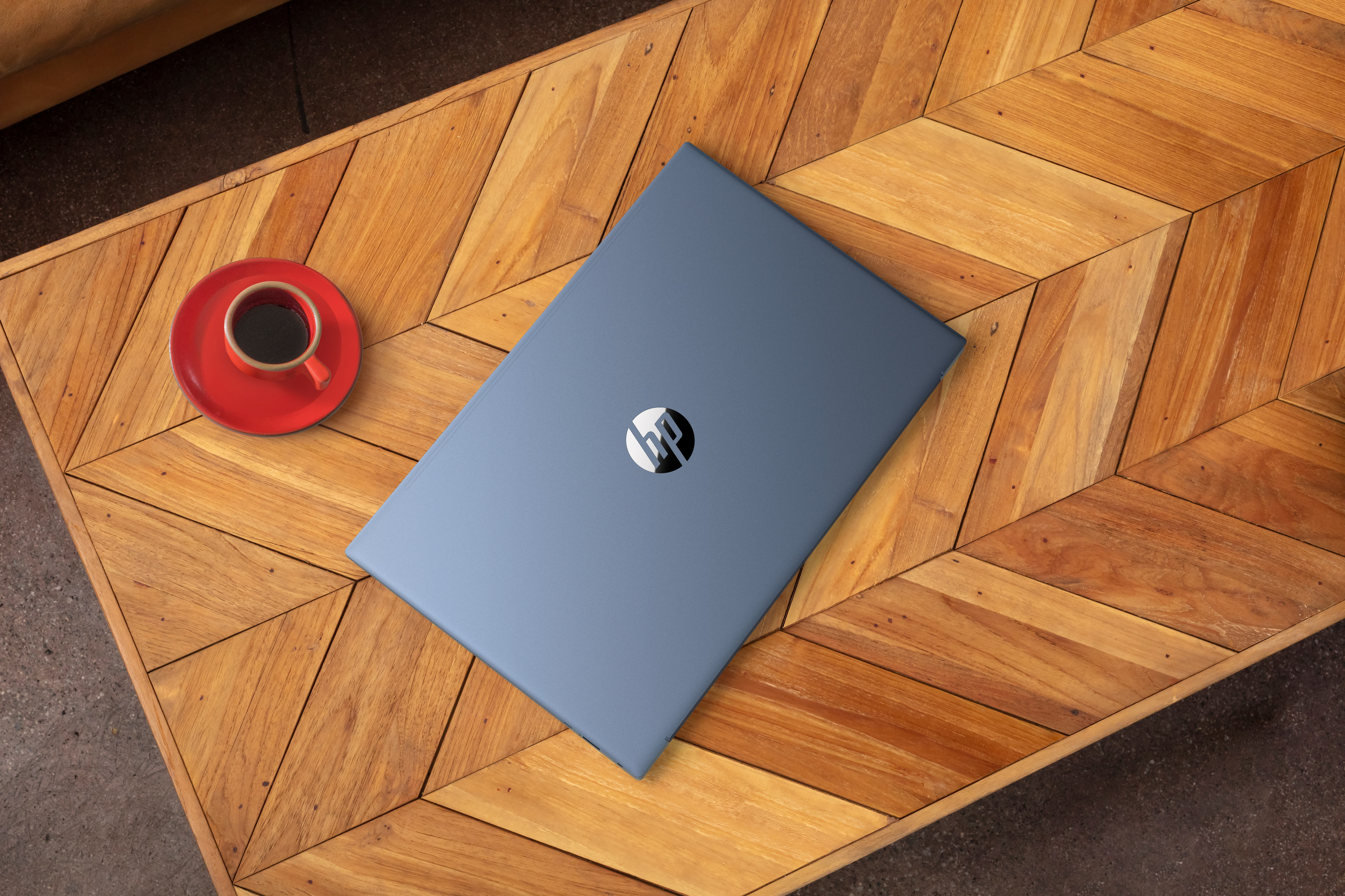Niebieski laptop z logo HP na drewnianym stole. Czerwony kubek z kawą obok.
