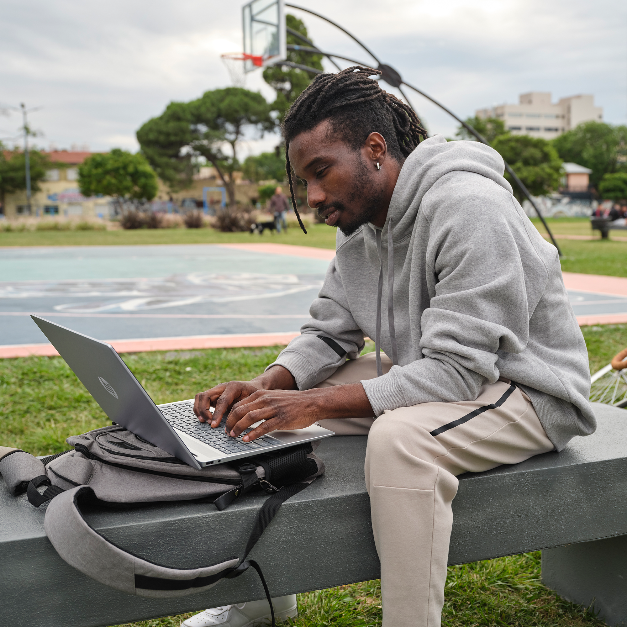Czarnoskóry mężczyzna w szarej bluzie z kapturem pracuje na laptopie na zewnątrz, na ławce. W tle boisko do koszykówki i drzewa.