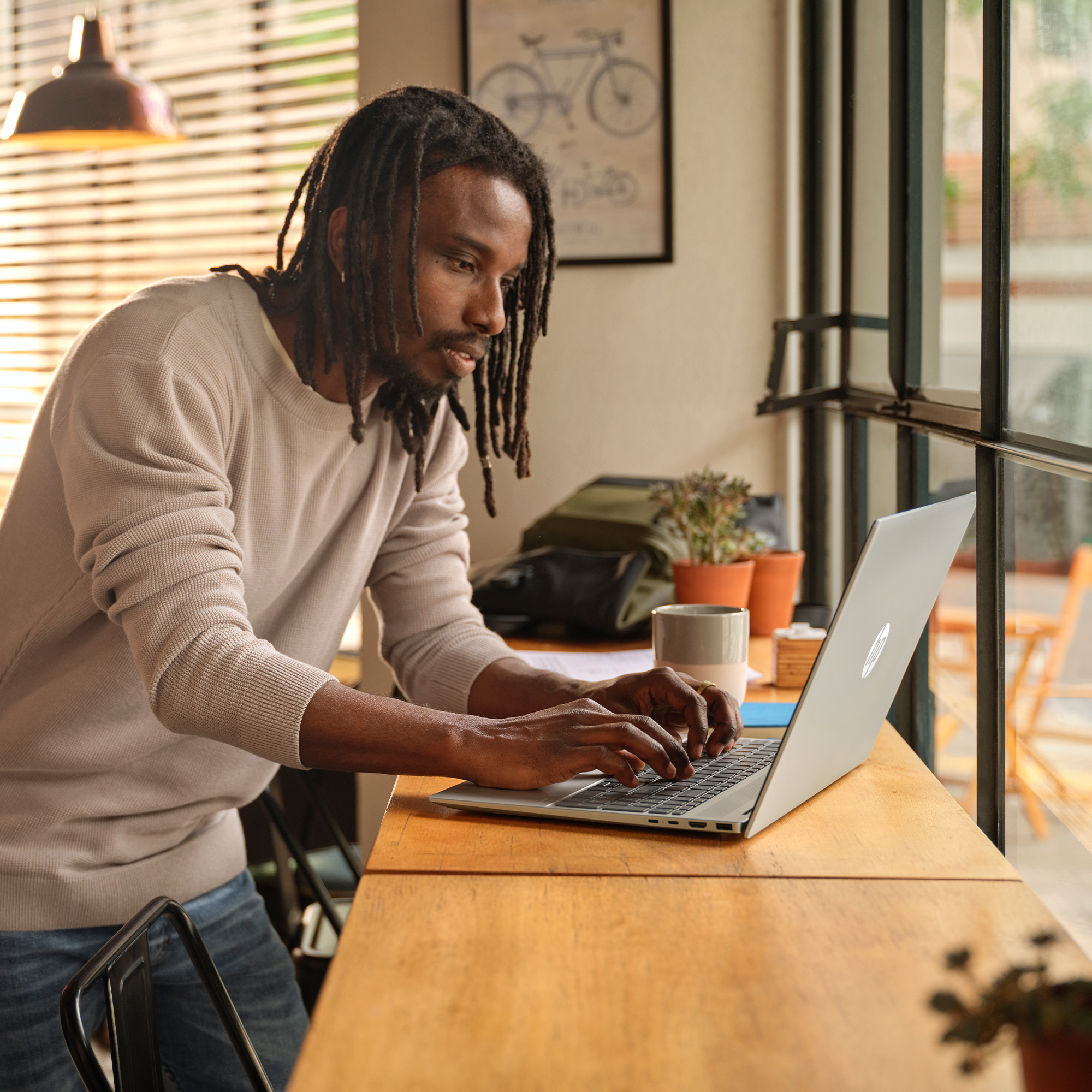 Czarnoskóry mężczyzna pisze na laptopie przy drewnianym stole w pobliżu okna. Na stole znajdują się również rośliny, kubek i plecak.