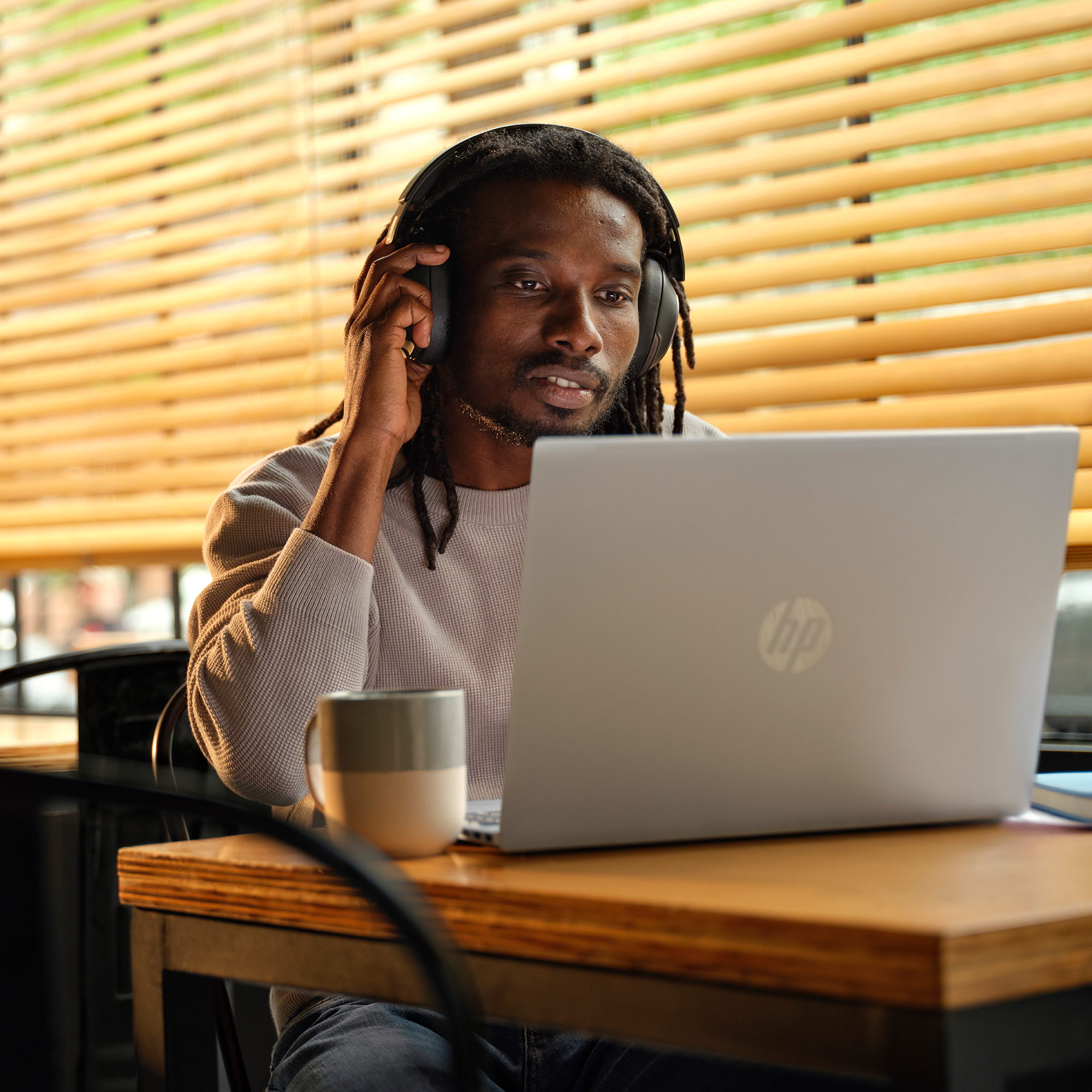 Czarny mężczyzna w słuchawkach korzysta z laptopa. Na stole stoi kubek. W tle żaluzje.