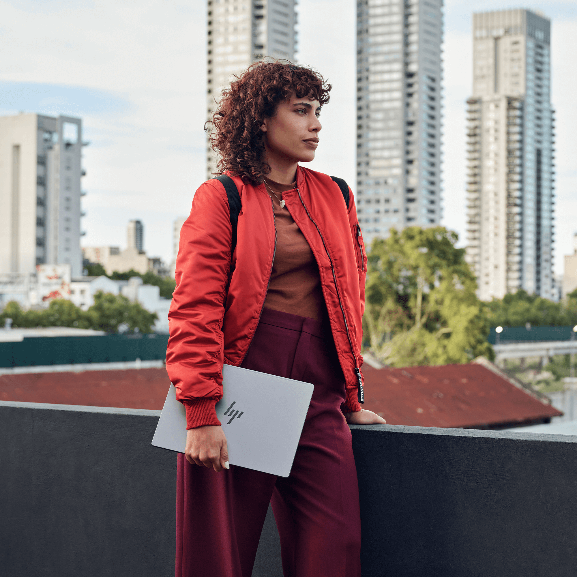 Kobieta w czerwonej kurtce trzyma laptopa na dachu. Budynki miejskie w tle. Odwraca wzrok.