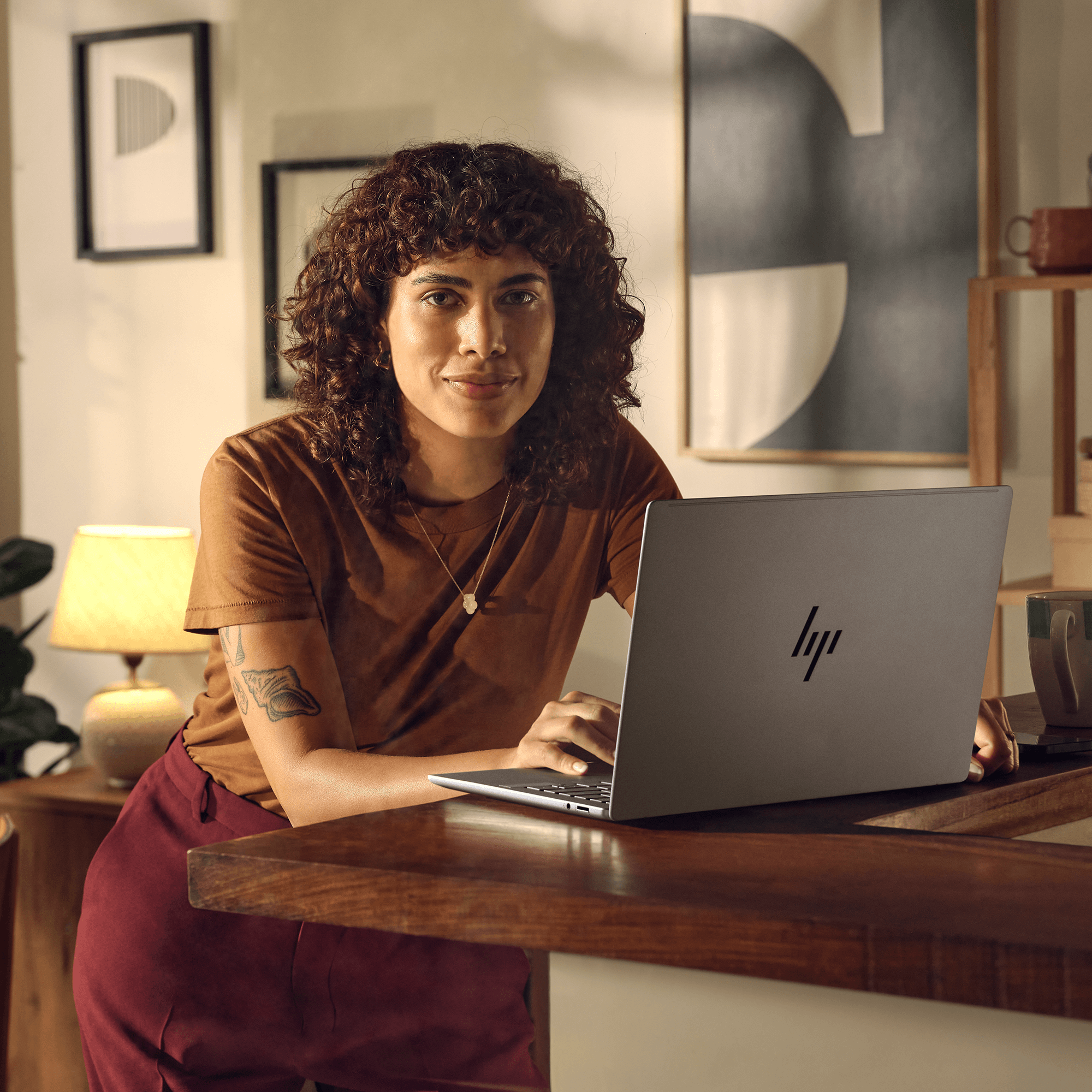 Kobieta z kręconymi włosami opiera się o blat, używając laptopa HP. Ma na sobie brązową koszulę i bordowe spodnie. W tle lampa.