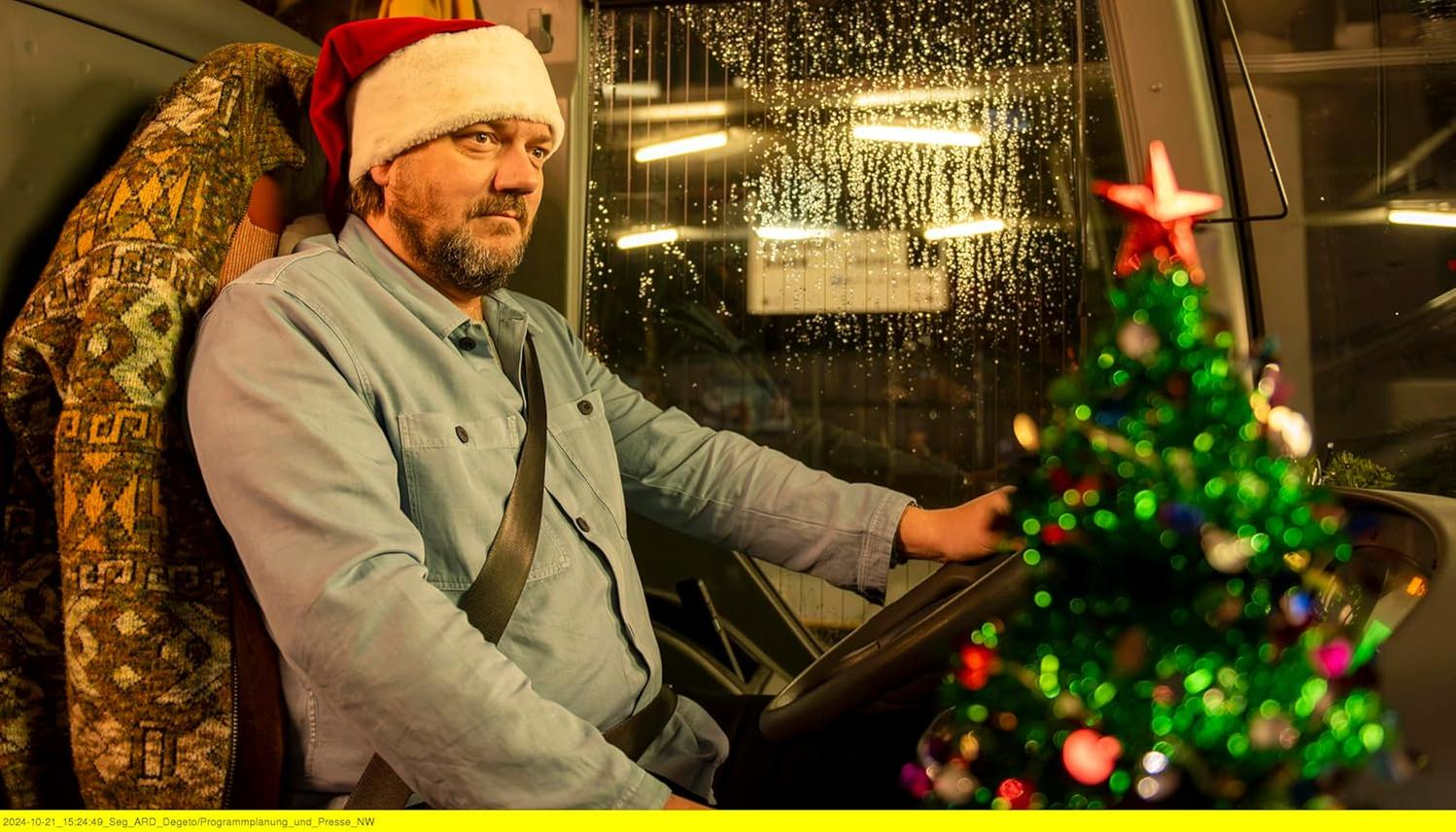 Mann mit Weihnachtsmütze fährt Bus, Weihnachtsbaum davor. Regen an der Scheibe. Pullover links.