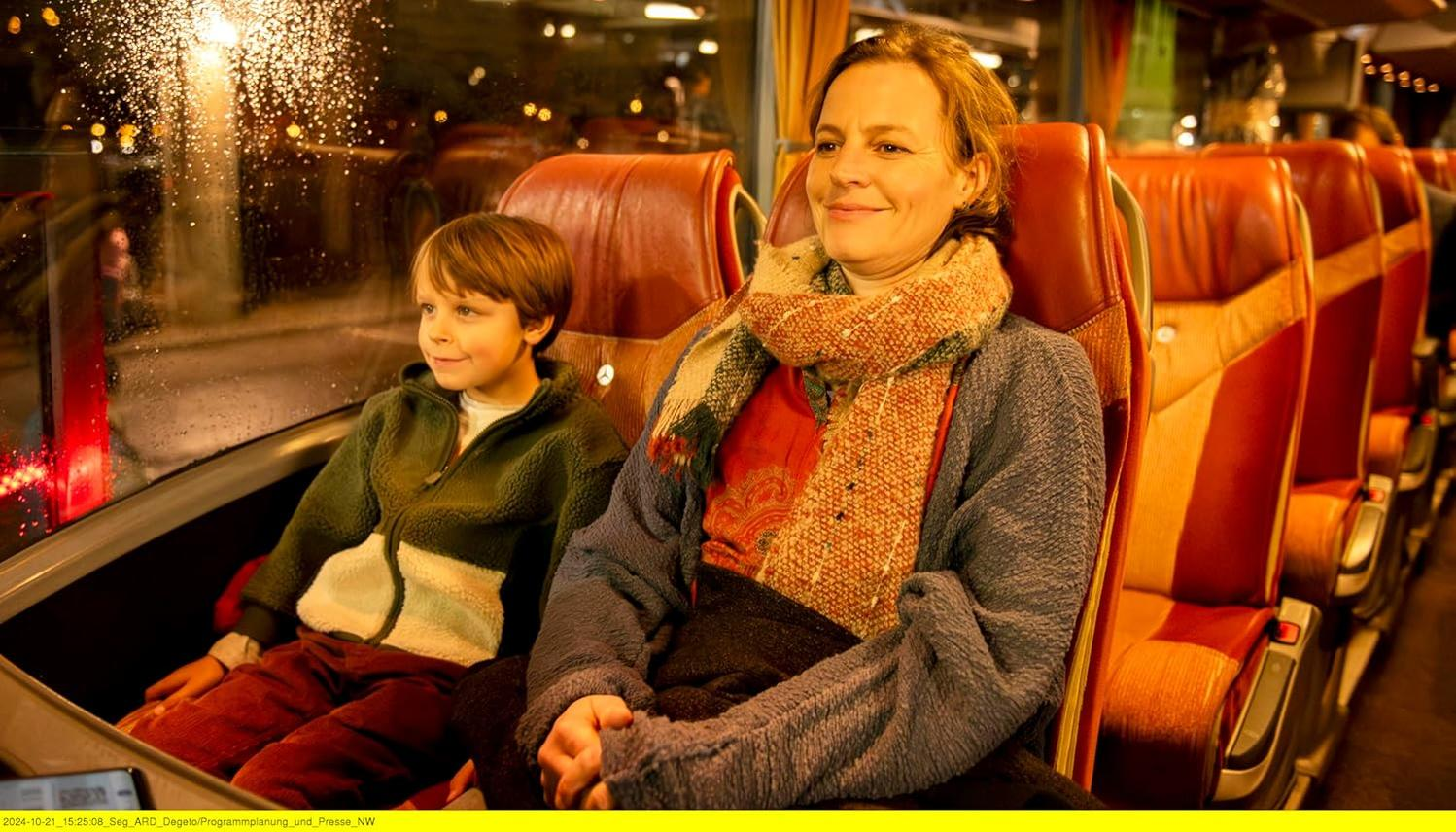 Eine Frau und ein Junge sitzen in einem Bus. Die Frau lächelt und trägt einen Schal. Der Junge schaut nachts aus dem Fenster.