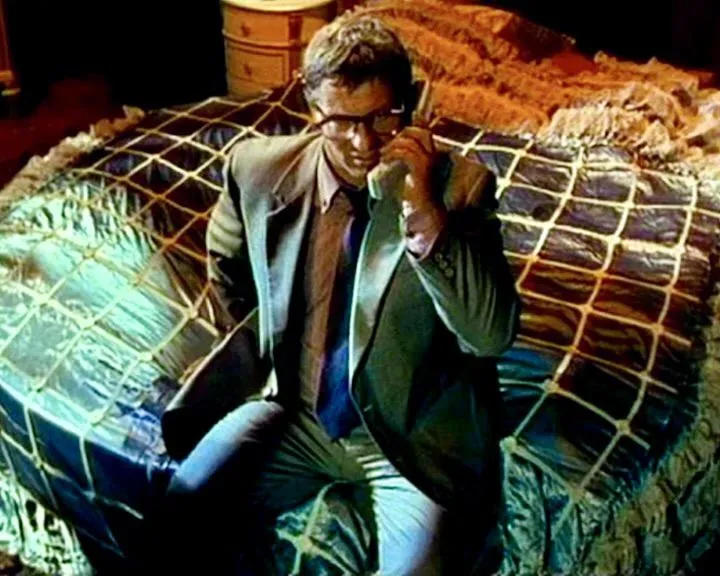 Ein Mann im Anzug mit Brille telefoniert, während er auf einem mit Netz bedeckten Objekt sitzt, vermutlich einem Bett.