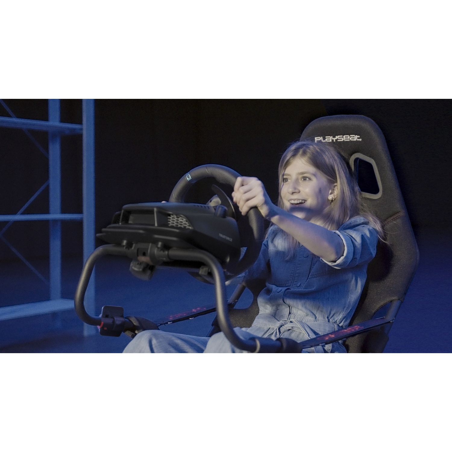Una ragazza sorride mentre tiene un volante in un simulatore di corse. È seduta su un sedile nero con il logo 'Playseat'.