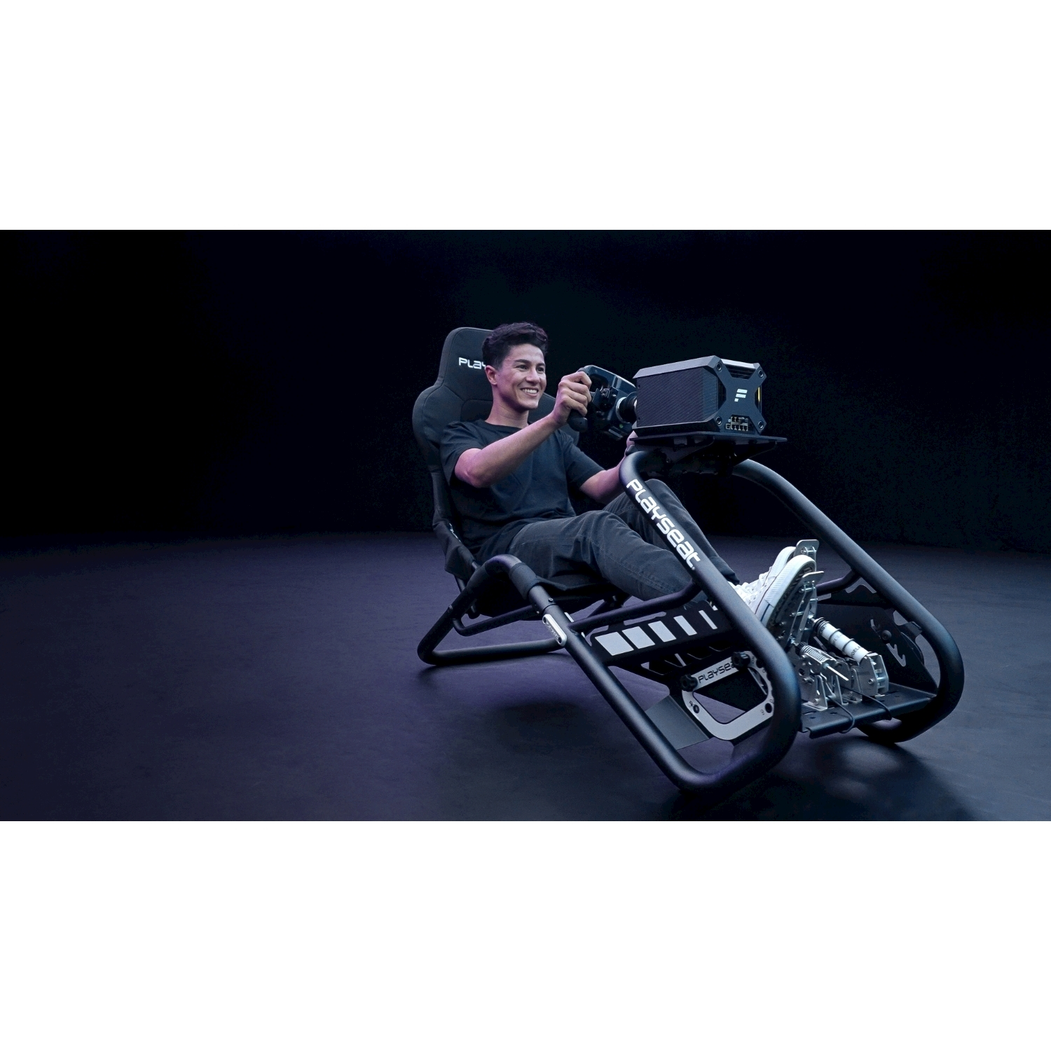 Un uomo sorride mentre usa un simulatore di corse. È seduto su una sedia PLAYSEAT, tenendo un volante. Lo sfondo è scuro.