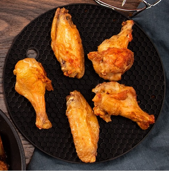 Sześć ugotowanych skrzydełek z kurczaka na czarnej macie o wzorze plastra miodu. Tło drewnianego stołu z ciemnoniebieską szmatką.