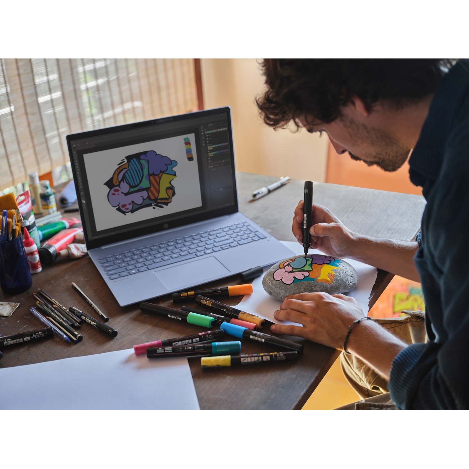 Un uomo dipinge un disegno colorato su una roccia. Un laptop mostra un'immagine simile. Pennarelli, penne e vernice sono sul tavolo.
