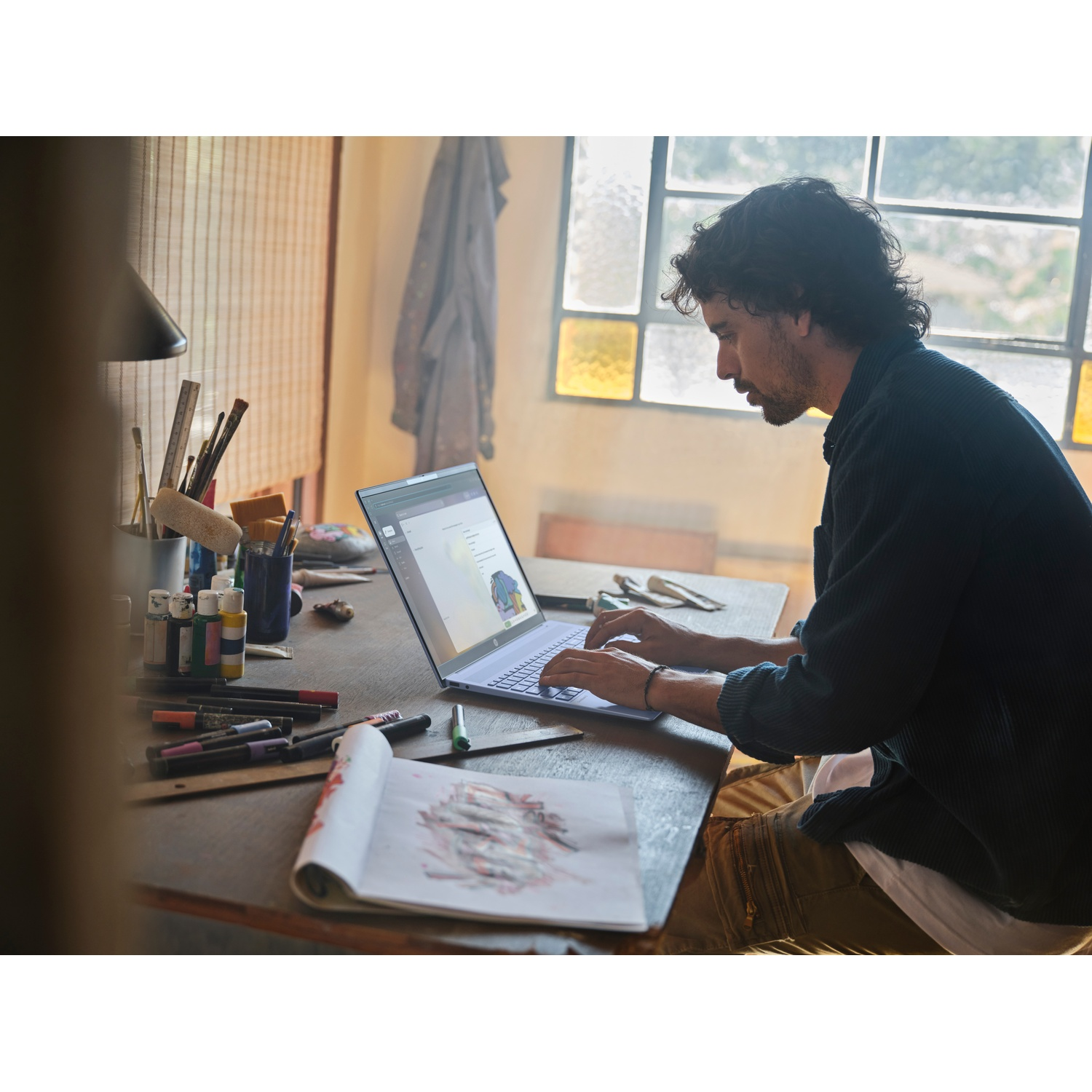 Un uomo scrive su un laptop a una scrivania di legno. Forniture artistiche e un blocco per schizzi sono vicini. Una finestra e un cappotto appeso sullo sfondo.