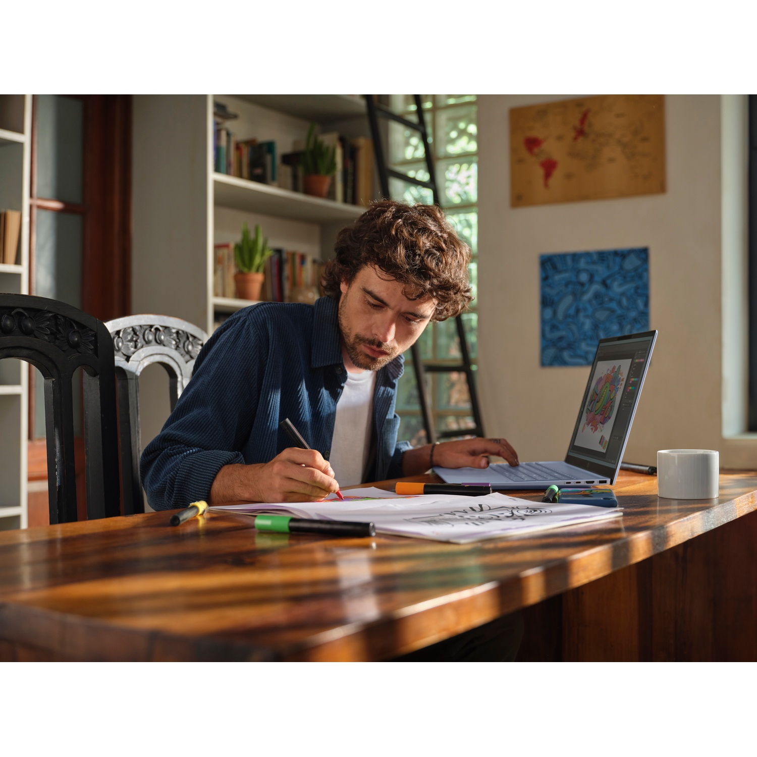 Un uomo disegna a un tavolo di legno con un laptop, pennarelli e una tazza. Librerie e arte murale sullo sfondo.