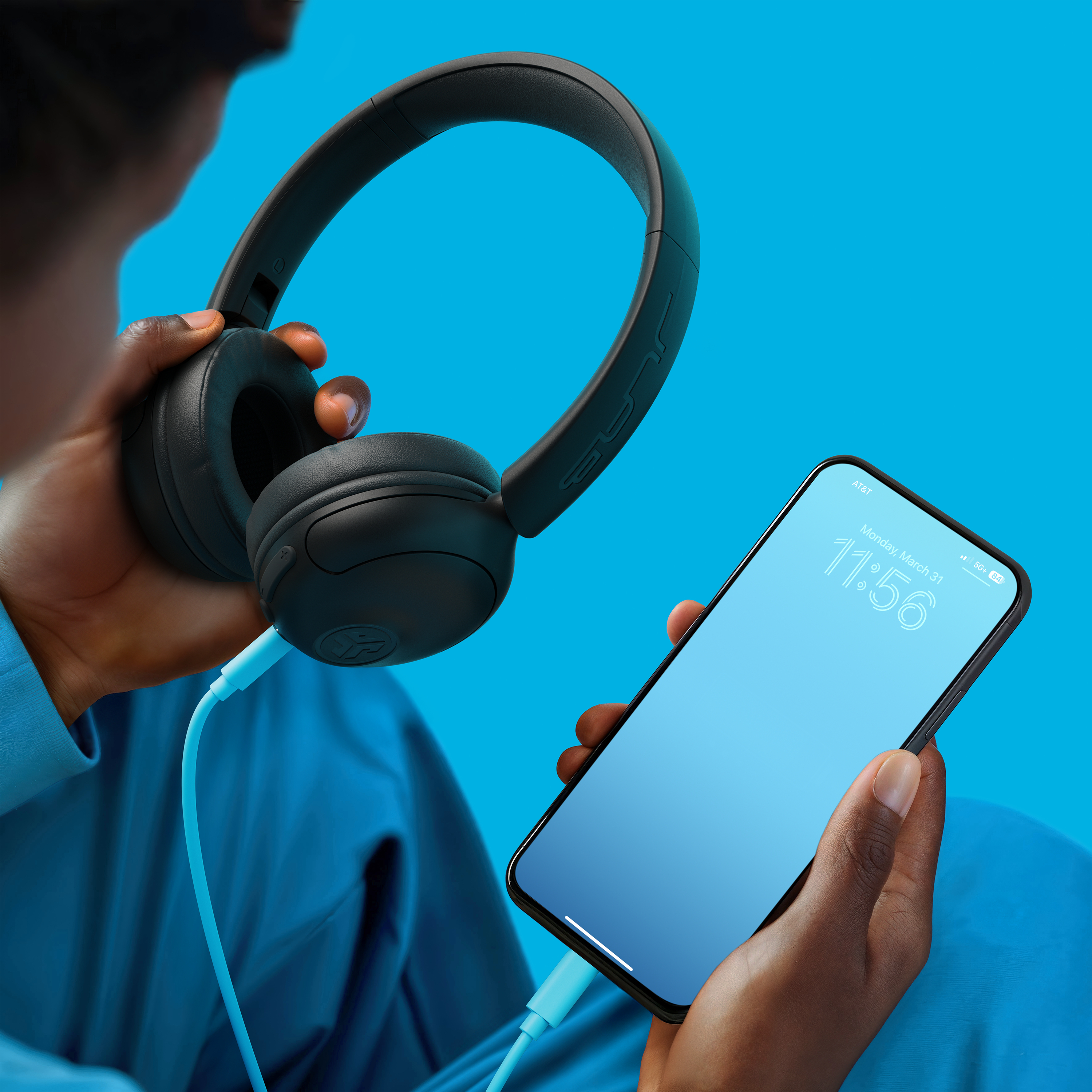 Eine Person hält schwarze Kopfhörer und ein Smartphone. Der Bildschirm des Telefons zeigt die Uhrzeit. Hellblauer Hintergrund.