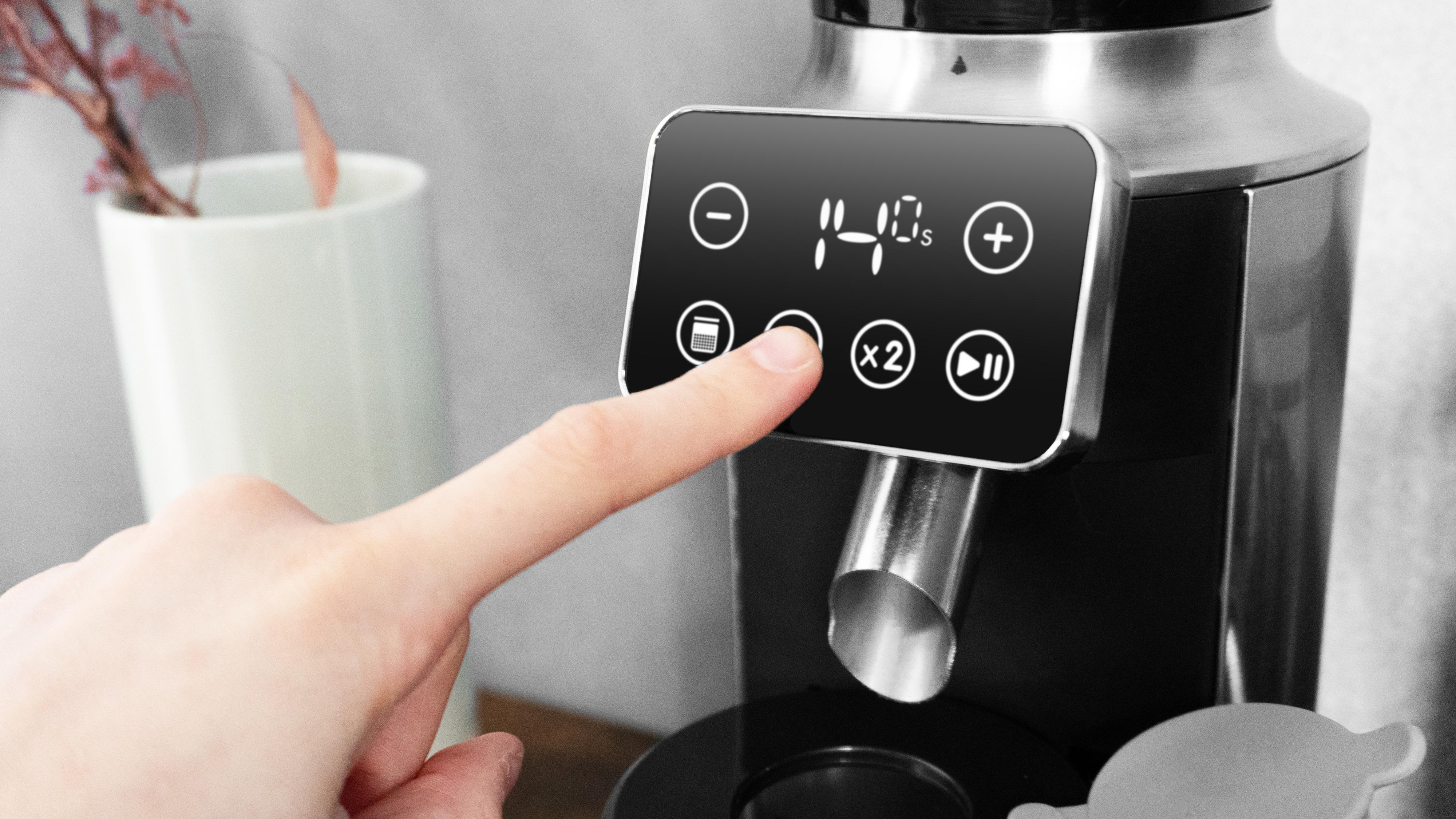 Eine Hand drückt eine Taste auf einer schwarzen Kaffeemaschine. Das Display zeigt '14s'. Eine weiße Tasse mit Blumen im Hintergrund.