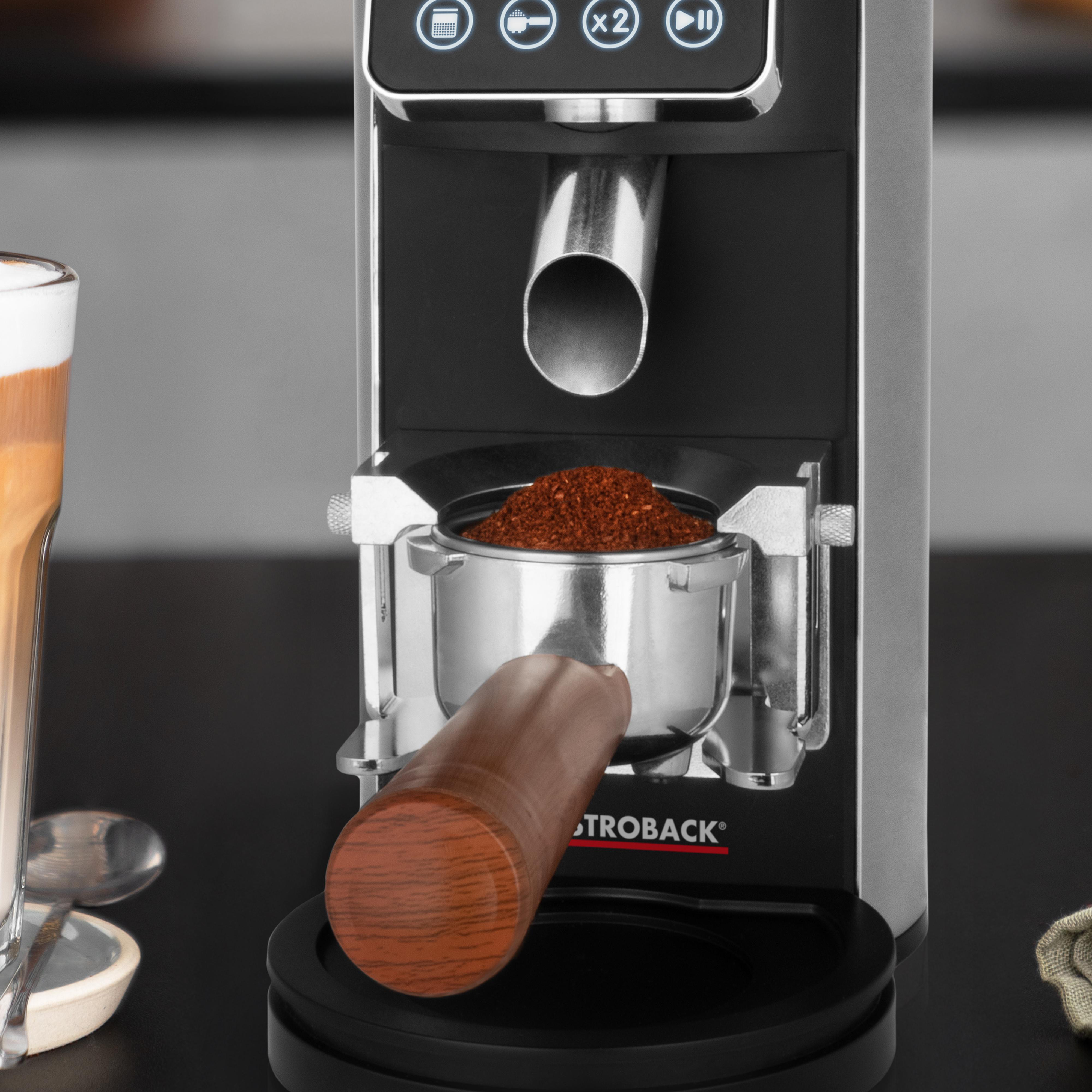 Kaffeemühle mit gefülltem Siebträger. Eine Kaffeetasse und ein Löffel sind auf dem Tisch.