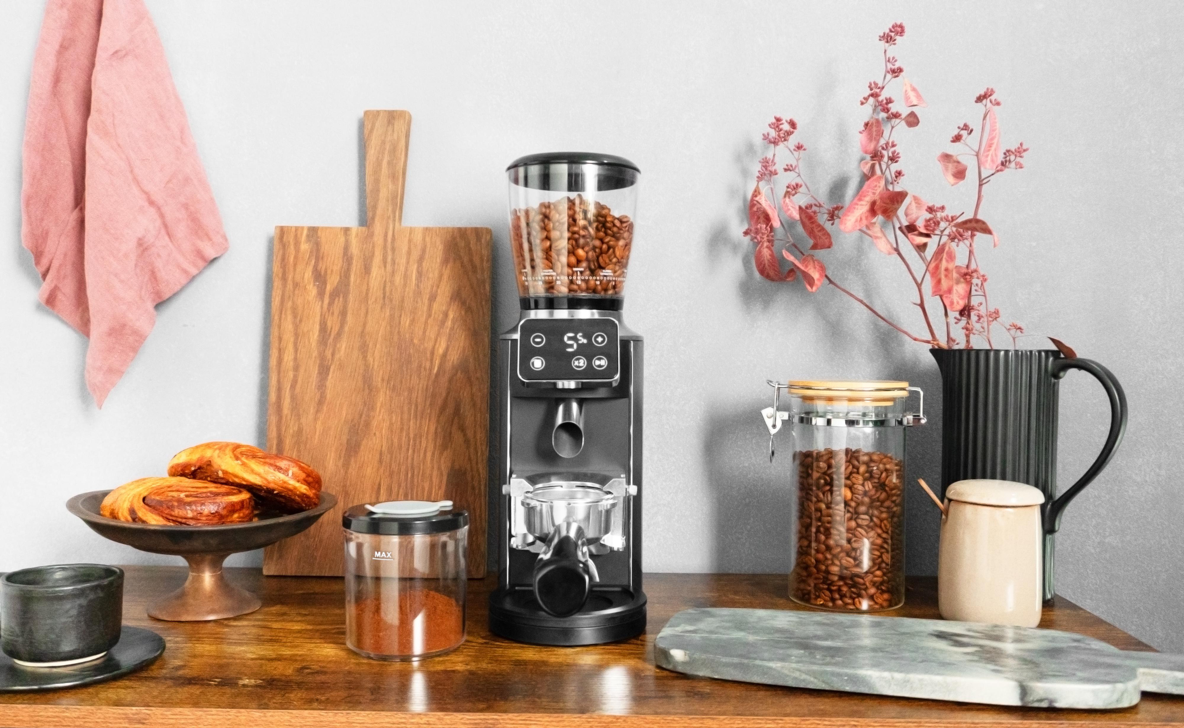 Kaffeemühle, Bohnen, Gebäck, Schneidebrett, rosa Tuch, Gläser und Blumen auf Holzoberfläche.