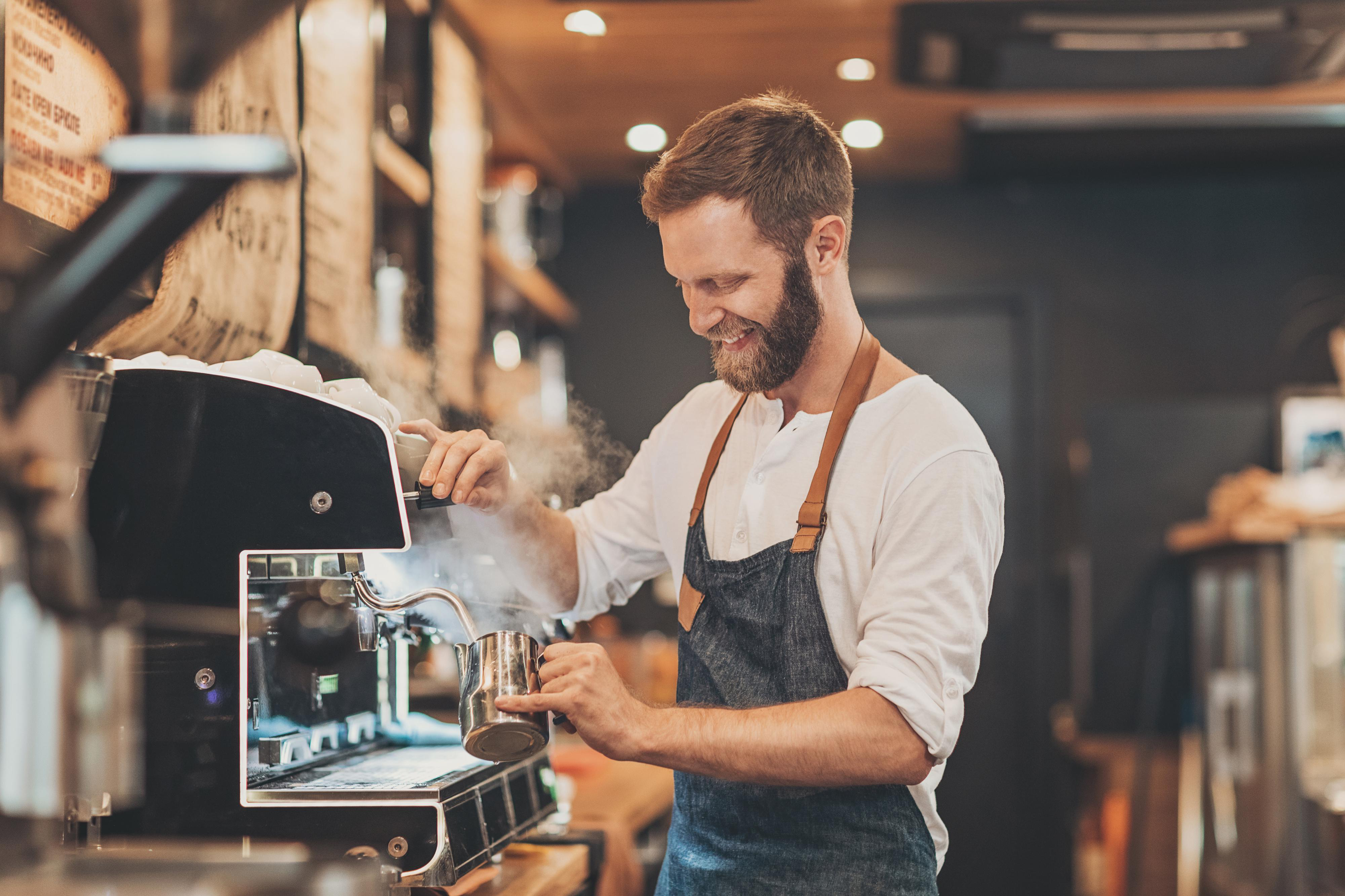 Ein Barista lächelt, während er Milch aufschäumt. Er trägt eine Schürze und ein weißes Hemd und arbeitet an einer Espressomaschine.