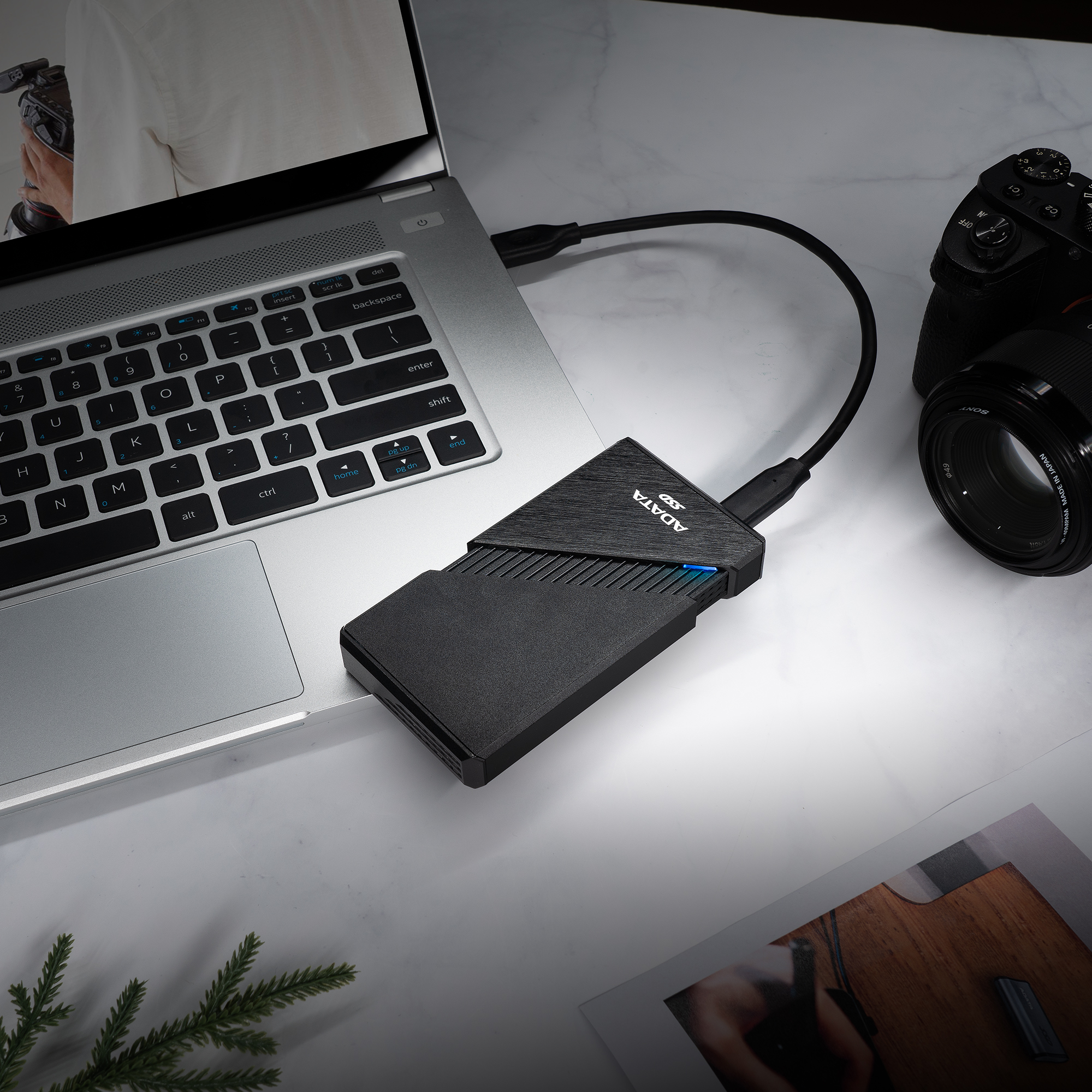 SSD ADATA nero collegato a un laptop. Fotocamera e foto su superficie bianca. Pianta verde visibile.