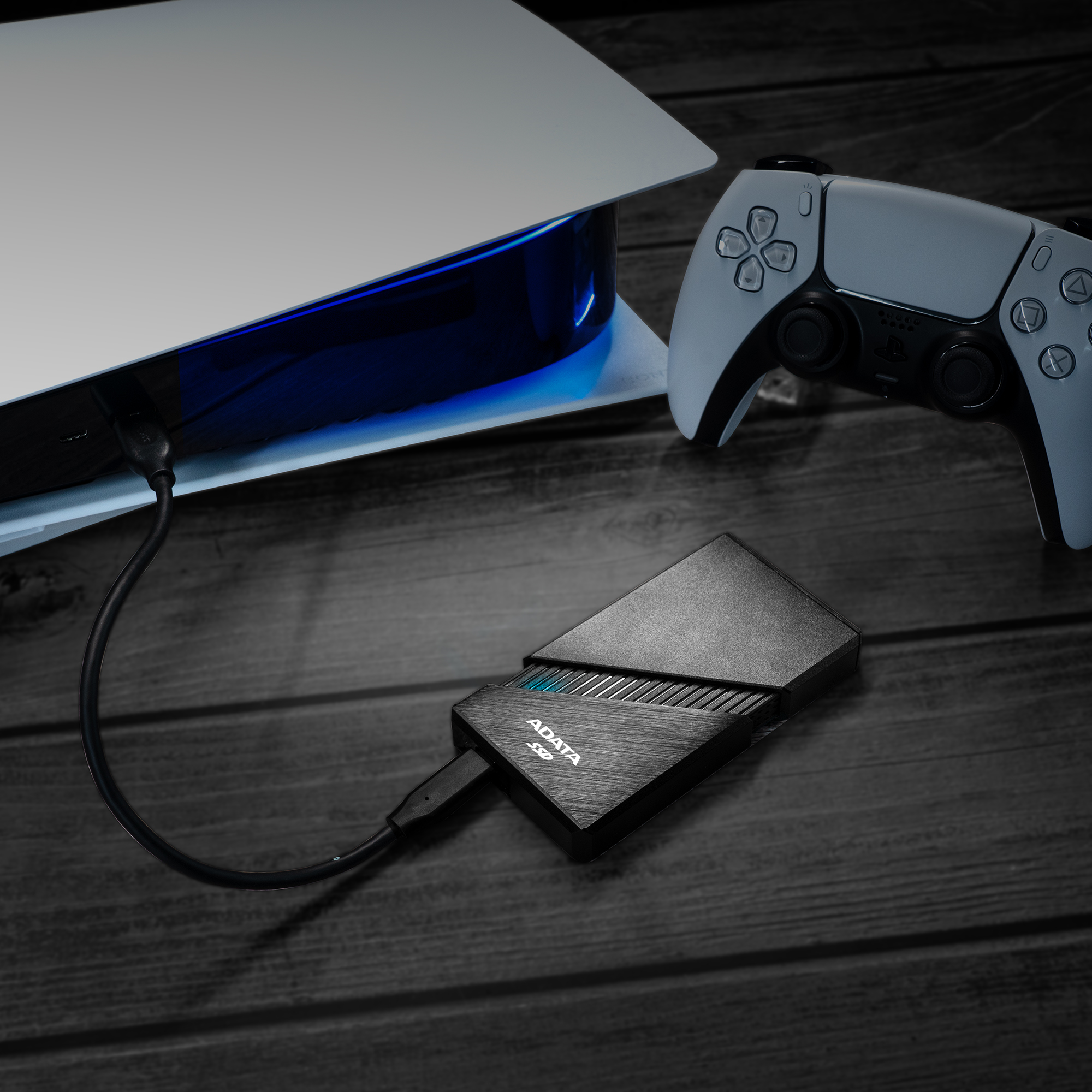 Console PS5, controller e SSD ADATA su superficie in legno. SSD collegato alla console. In scala di grigi.