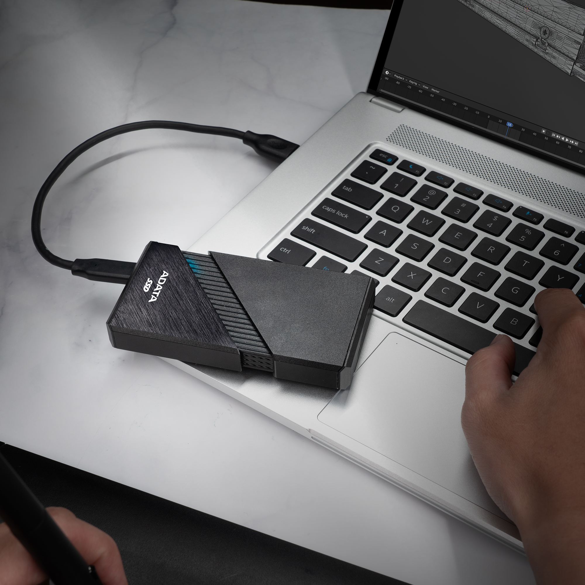 SSD ADATA nera collegata a un laptop. Una mano è sulla tastiera, lo schermo mostra un wireframe.
