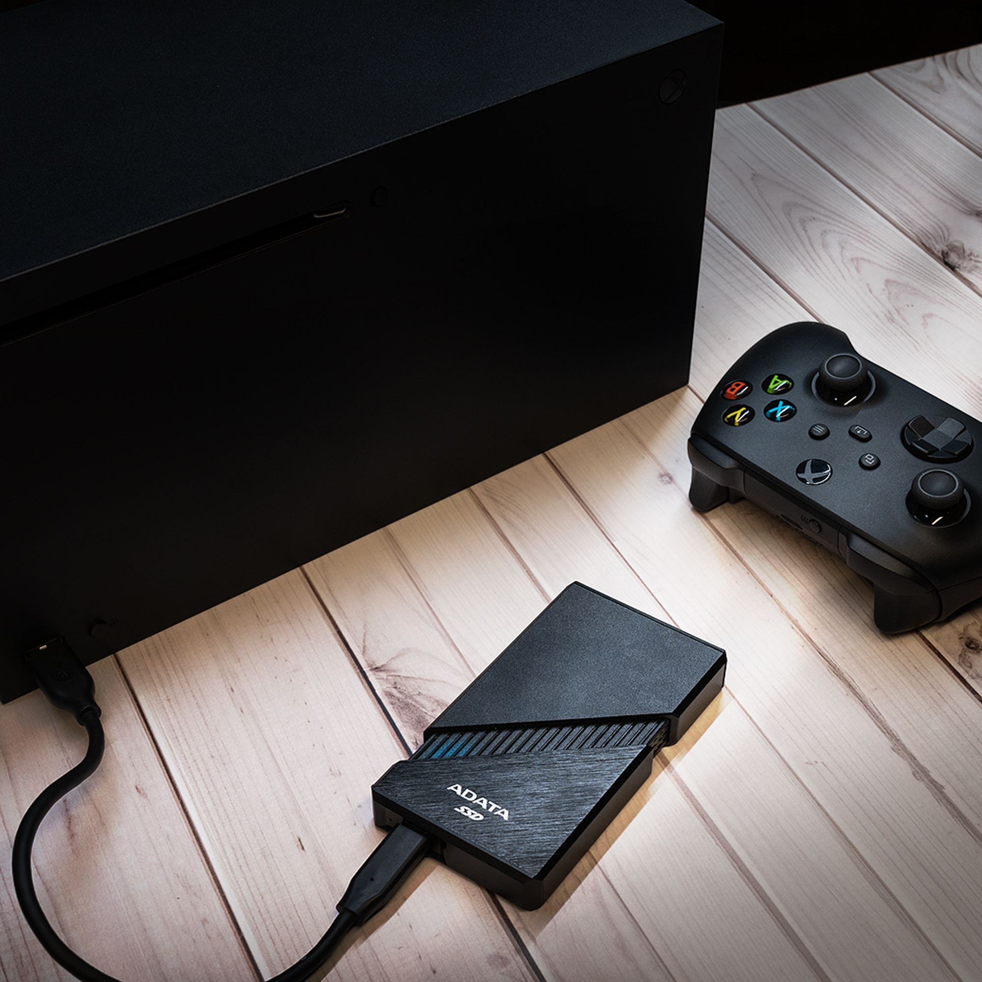 Console di gioco, controller e SSD esterno neri su superficie in legno. L'SSD è collegato alla console. Controller a destra.