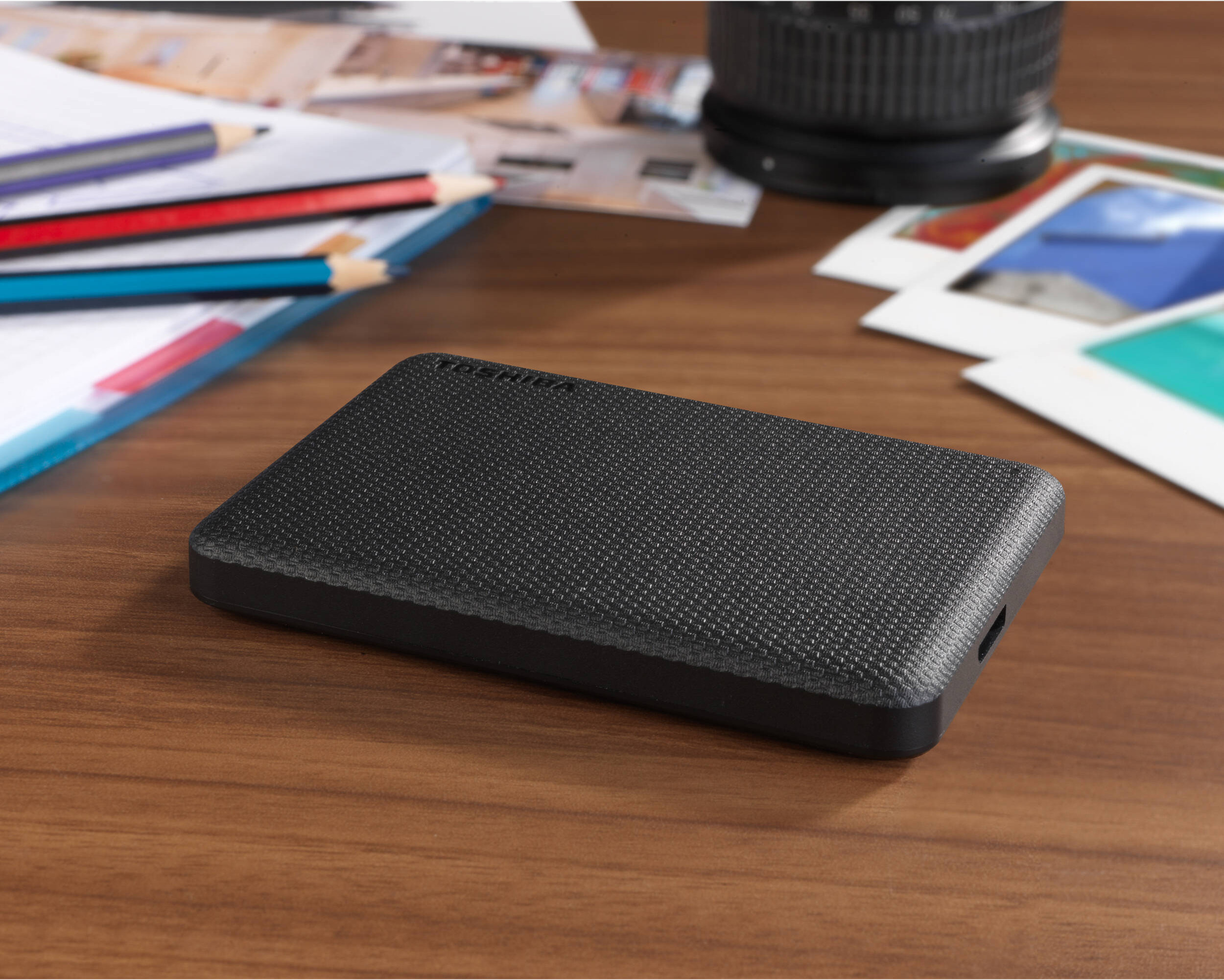 Disque dur externe noir sur un bureau en bois avec crayons, objectif et photos. Le disque est mis au point.