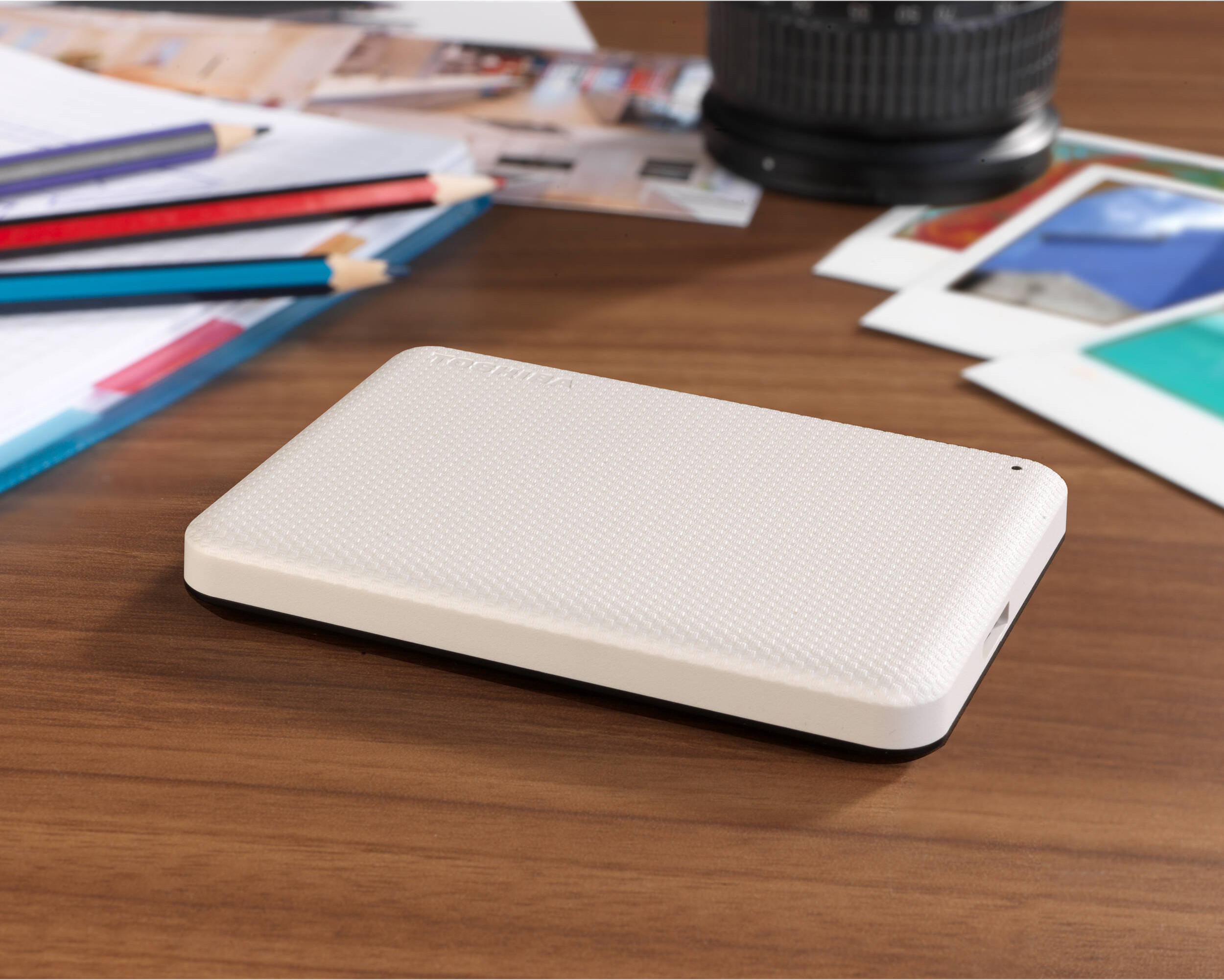 Un disque dur externe crème est posé sur un bureau en bois. Crayon, papier, objectif et photos en arrière-plan.