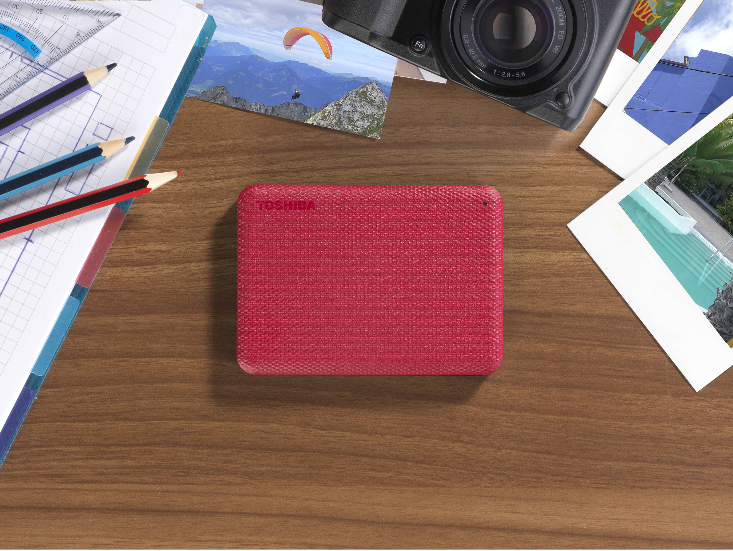 Disque dur externe rouge Toshiba sur table en bois, entouré de photos, crayons et appareil photo.