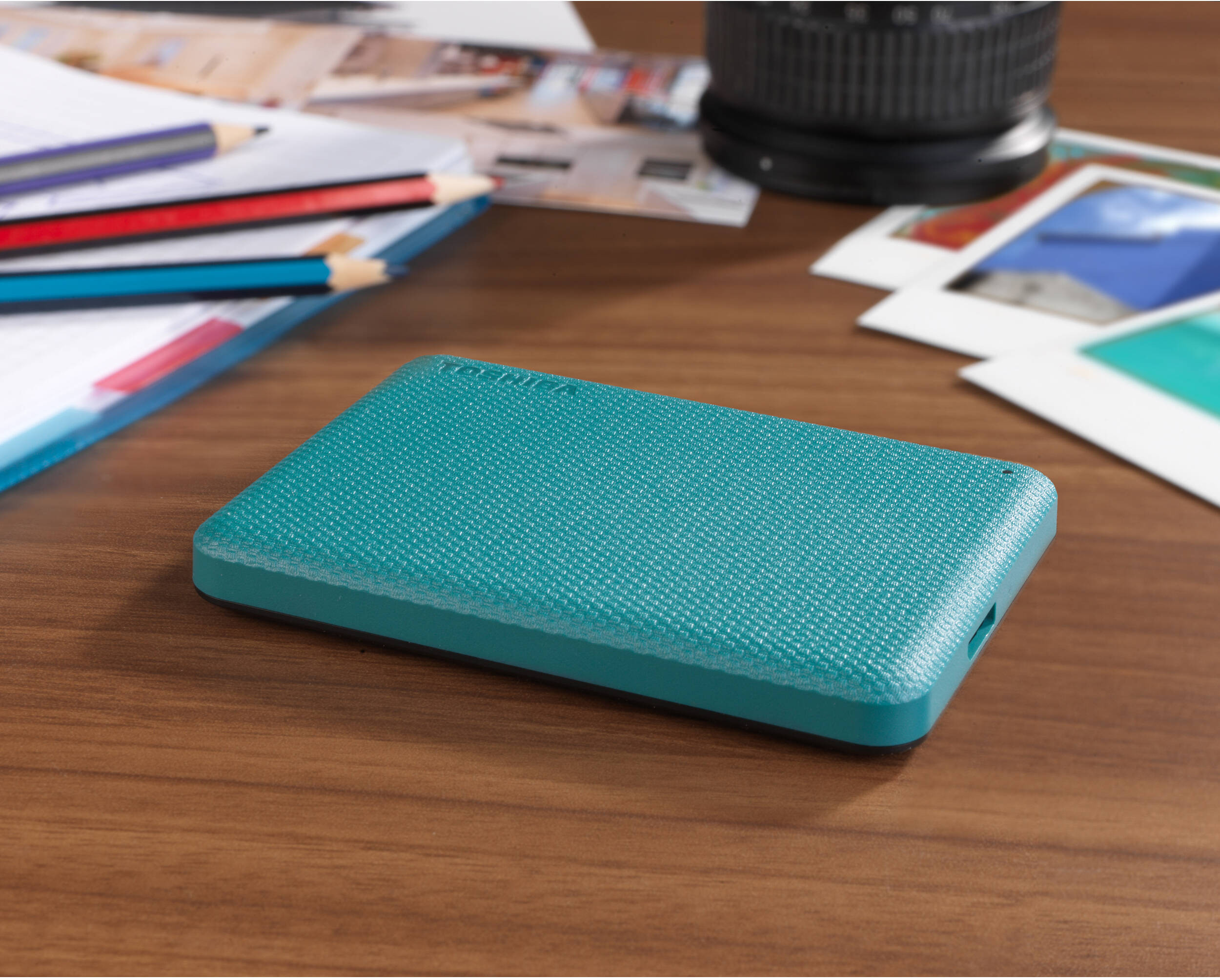 Un disque dur externe turquoise est posé sur une table en bois, avec des crayons, des photos et un objectif d'appareil photo en arrière-plan.