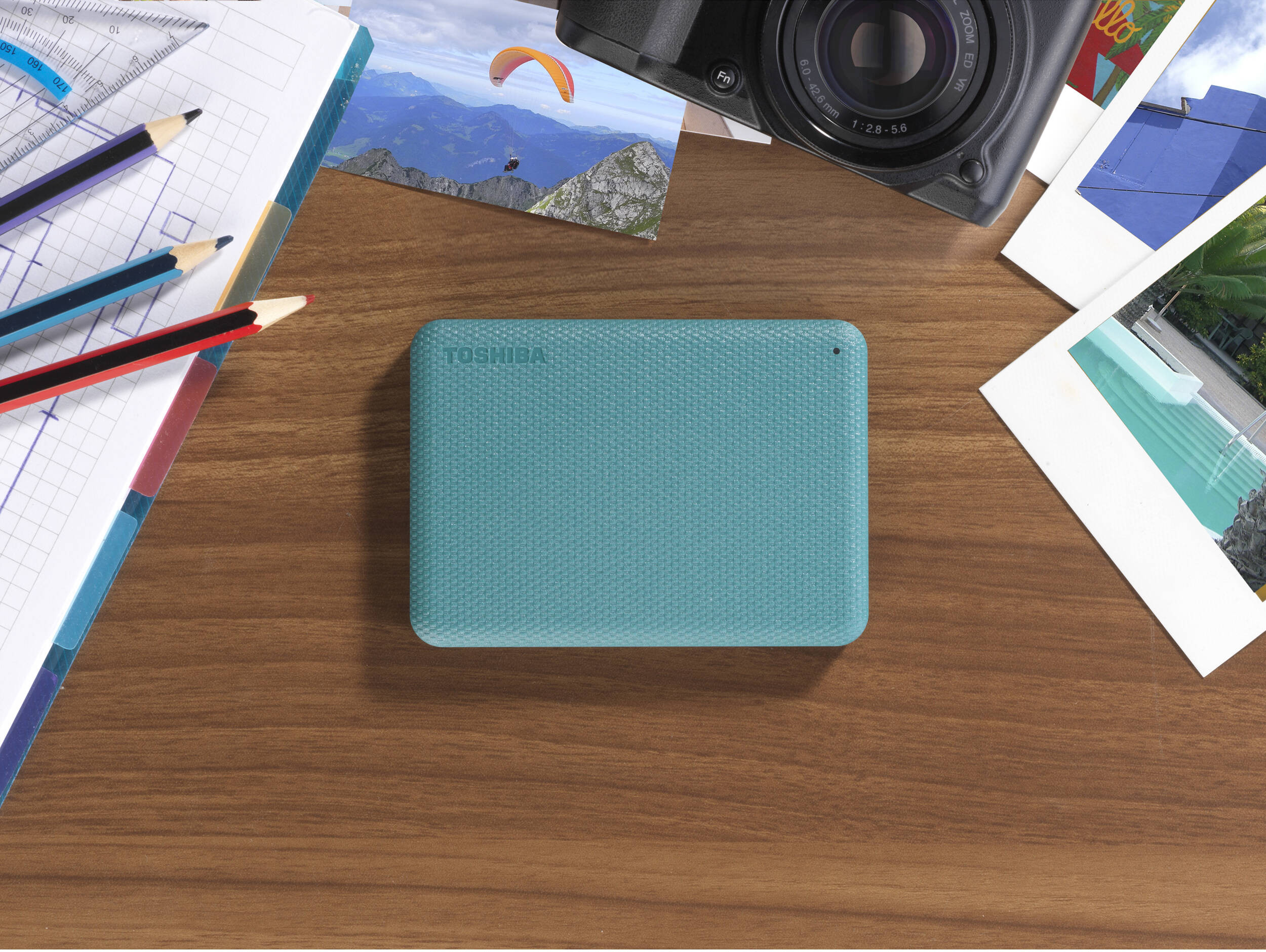 Un disque dur Toshiba turquoise sur une surface en bois, entouré de photos, d'un appareil photo, de crayons et d'une règle.