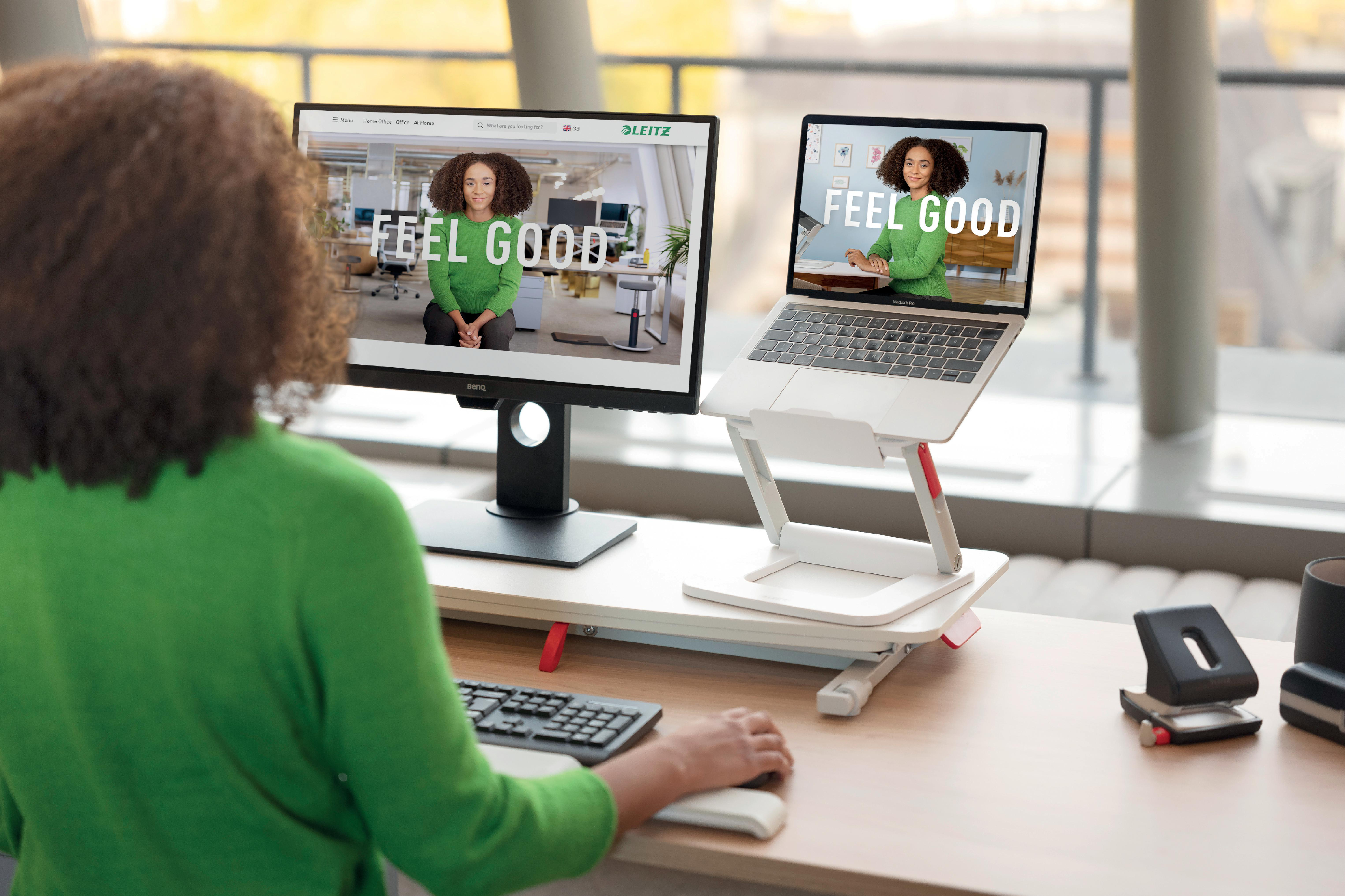 Eine Person arbeitet an einem Schreibtisch an einem Computer und Laptop, beide zeigen 'FEEL GOOD'.