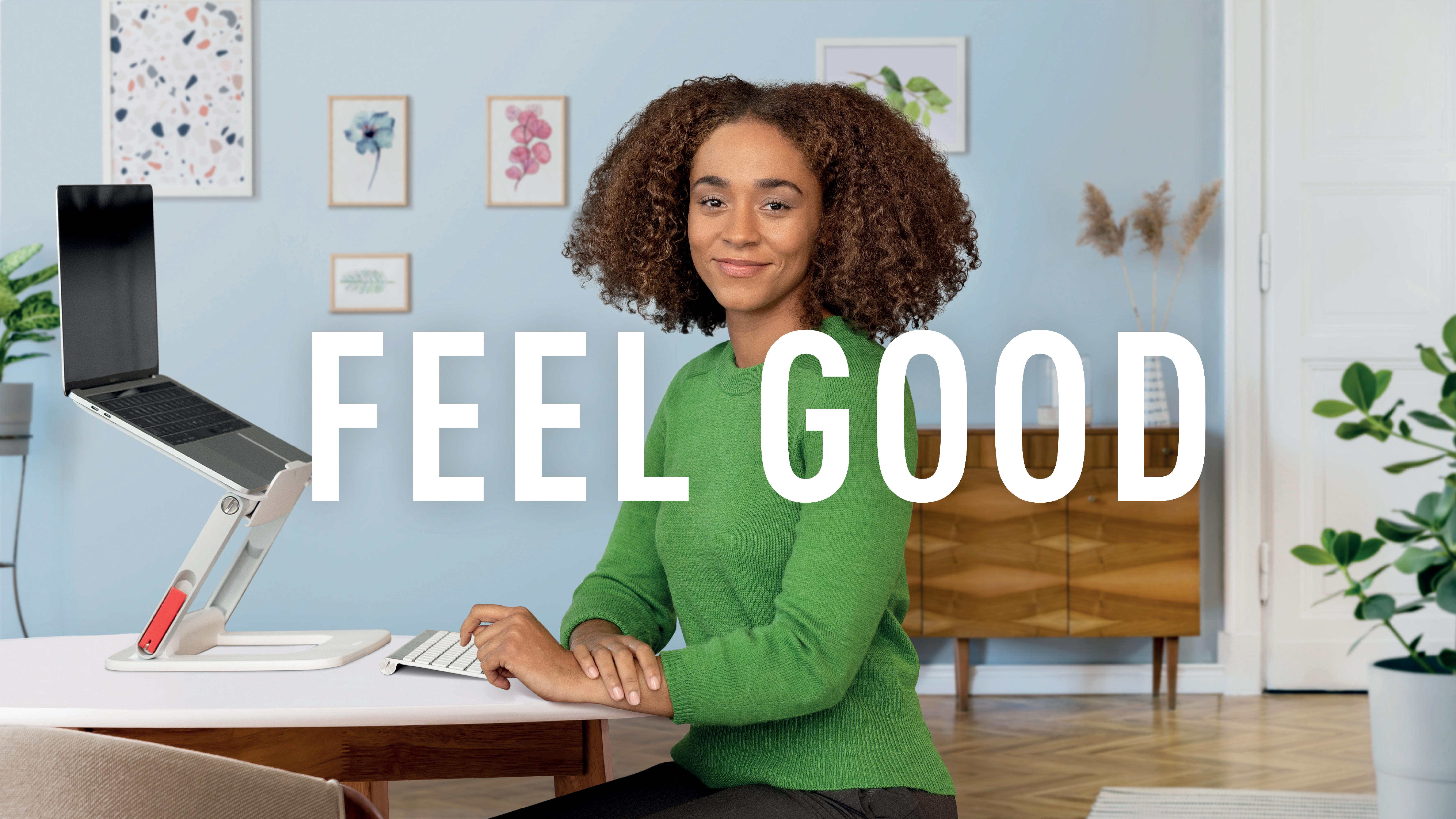 Eine Frau in einem grünen Pullover sitzt an einem Schreibtisch mit einem Laptop. Das Wort FEEL GOOD steht in weißen Buchstaben.