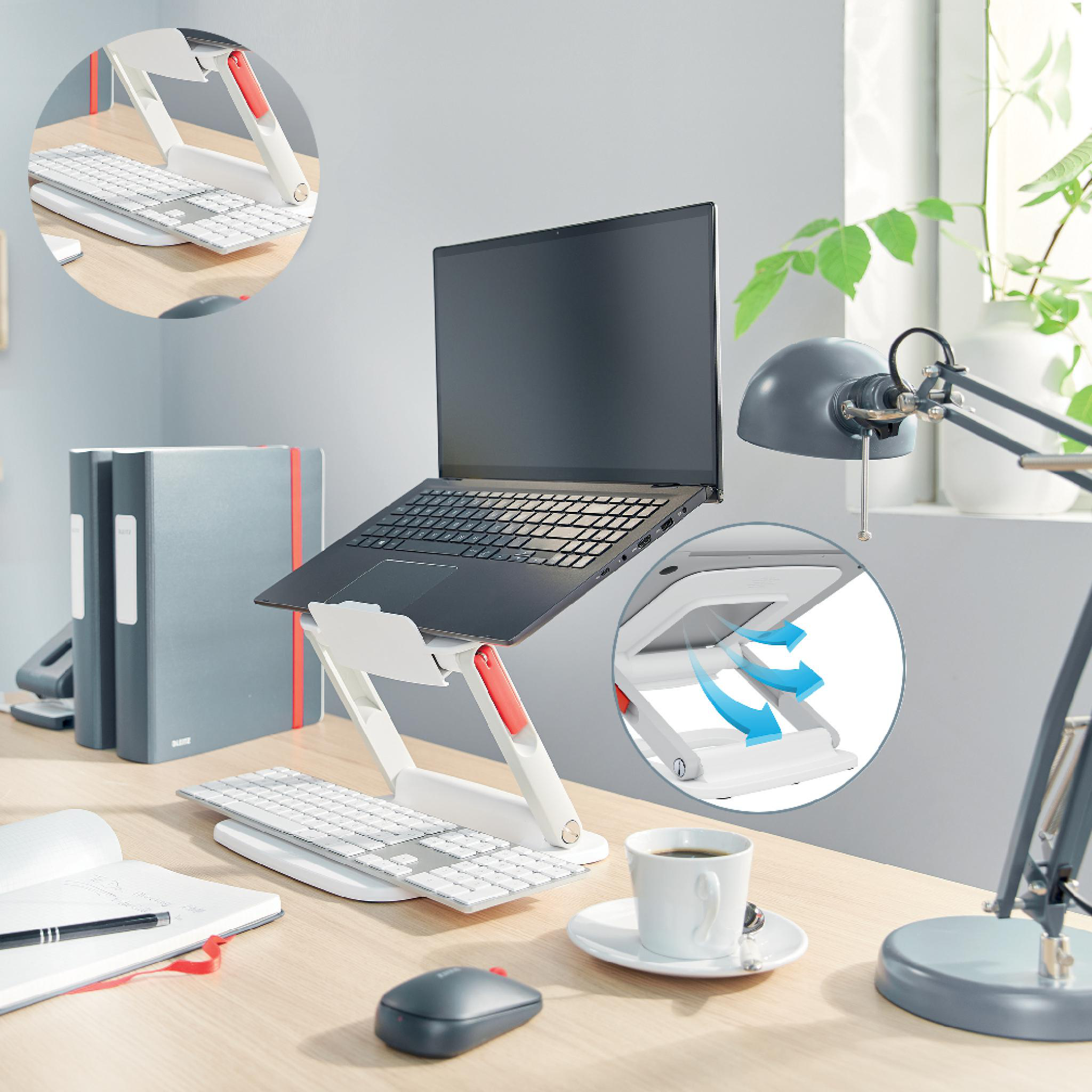 Laptop auf weißem Ständer, Tastatur, Maus und Kaffeetasse auf einem Holztisch. Eine Lampe, Ordner und eine Pflanze sind sichtbar.