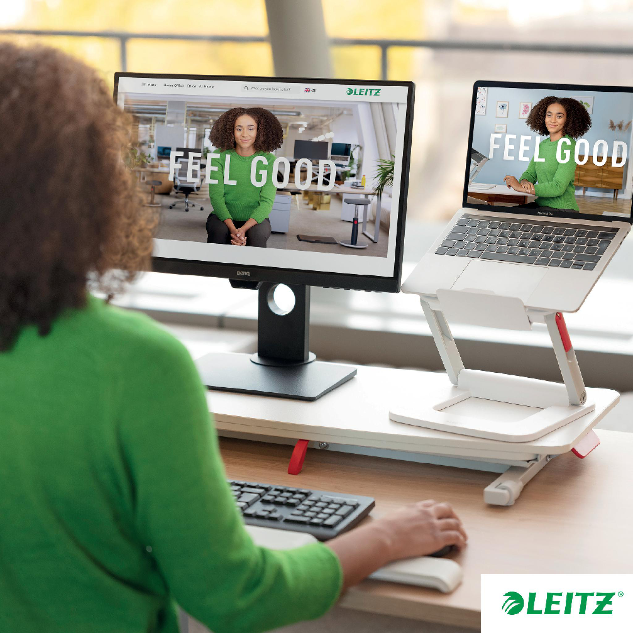 Frau am Schreibtisch mit zwei Monitoren, einer mit 'FEEL GOOD'-Text. Laptop auf Ständer. Grüner Pullover. LEITZ-Logo in der Ecke.