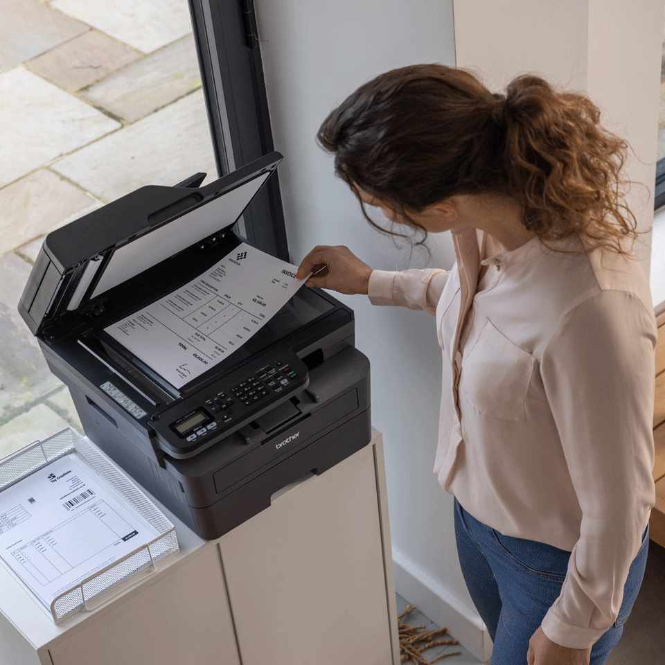 Eine Frau legt eine Rechnung in einen Brother-Drucker. Der Drucker steht auf einem weißen Schrank neben einem Fenster. Ein Drahtgestell hält Papiere.