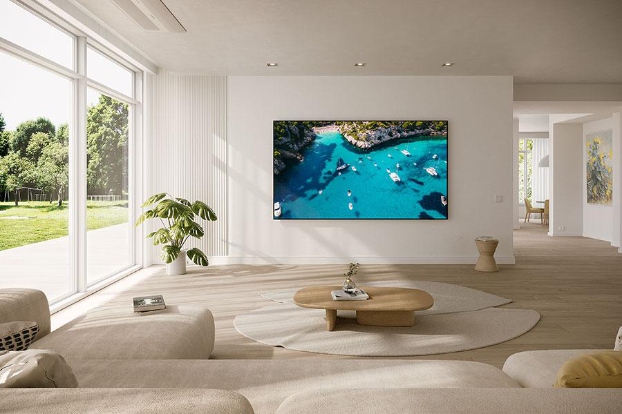 Wohnzimmer mit großem Fernseher, der eine Strandszene zeigt. Eine Couch, ein Couchtisch und ein großes Fenster sind sichtbar.