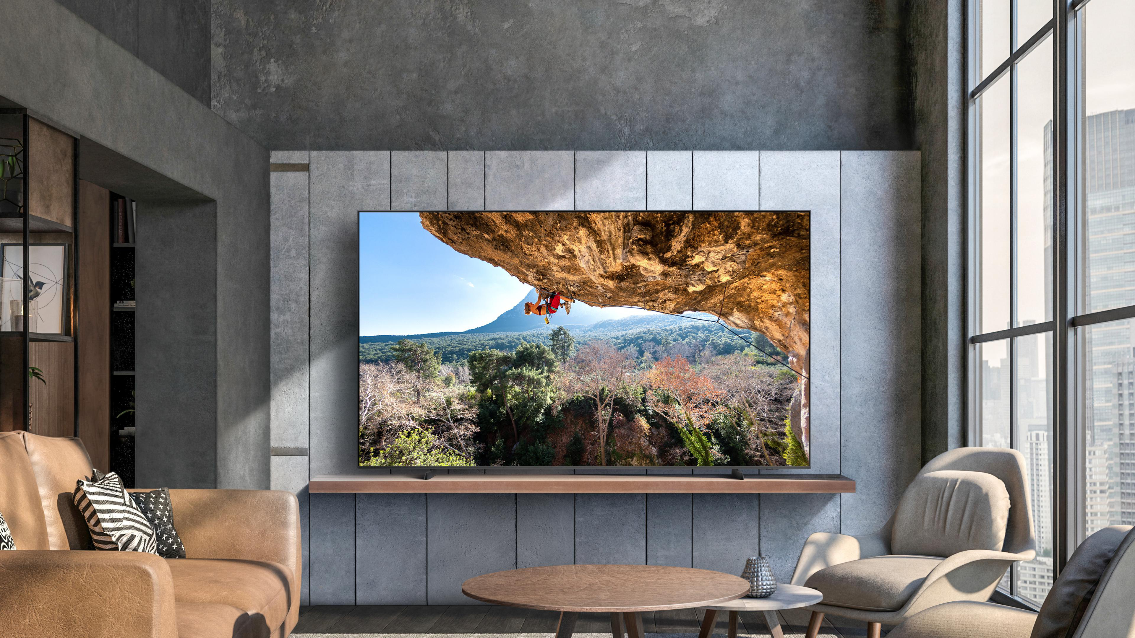 Wohnzimmer mit TV, der einen Kletterer zeigt. Braunes Ledersofa, runder Tisch und Stadtblick.
