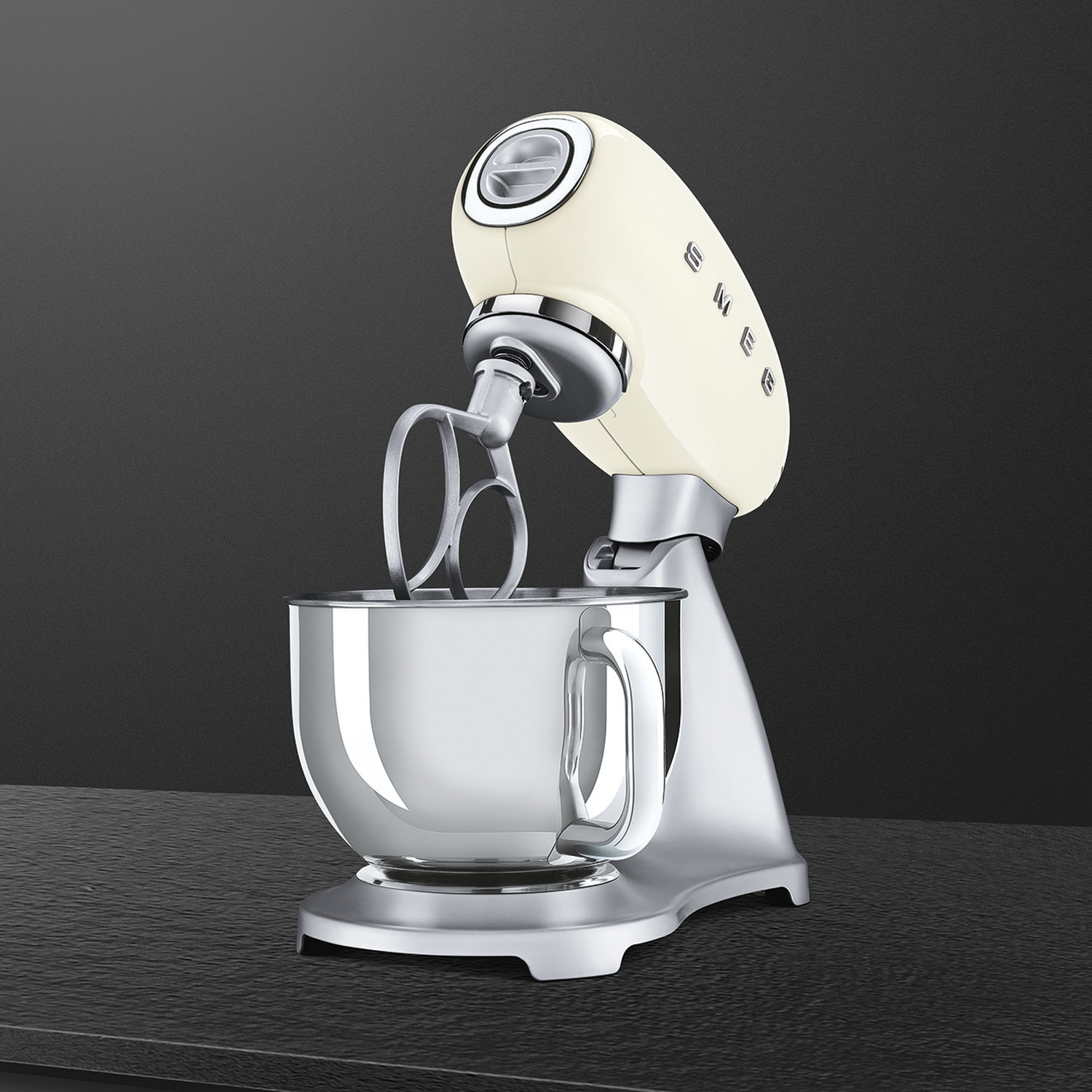 A cream-colored stand mixer with a silver bowl and attachments sits on a dark surface. The background is dark gray.