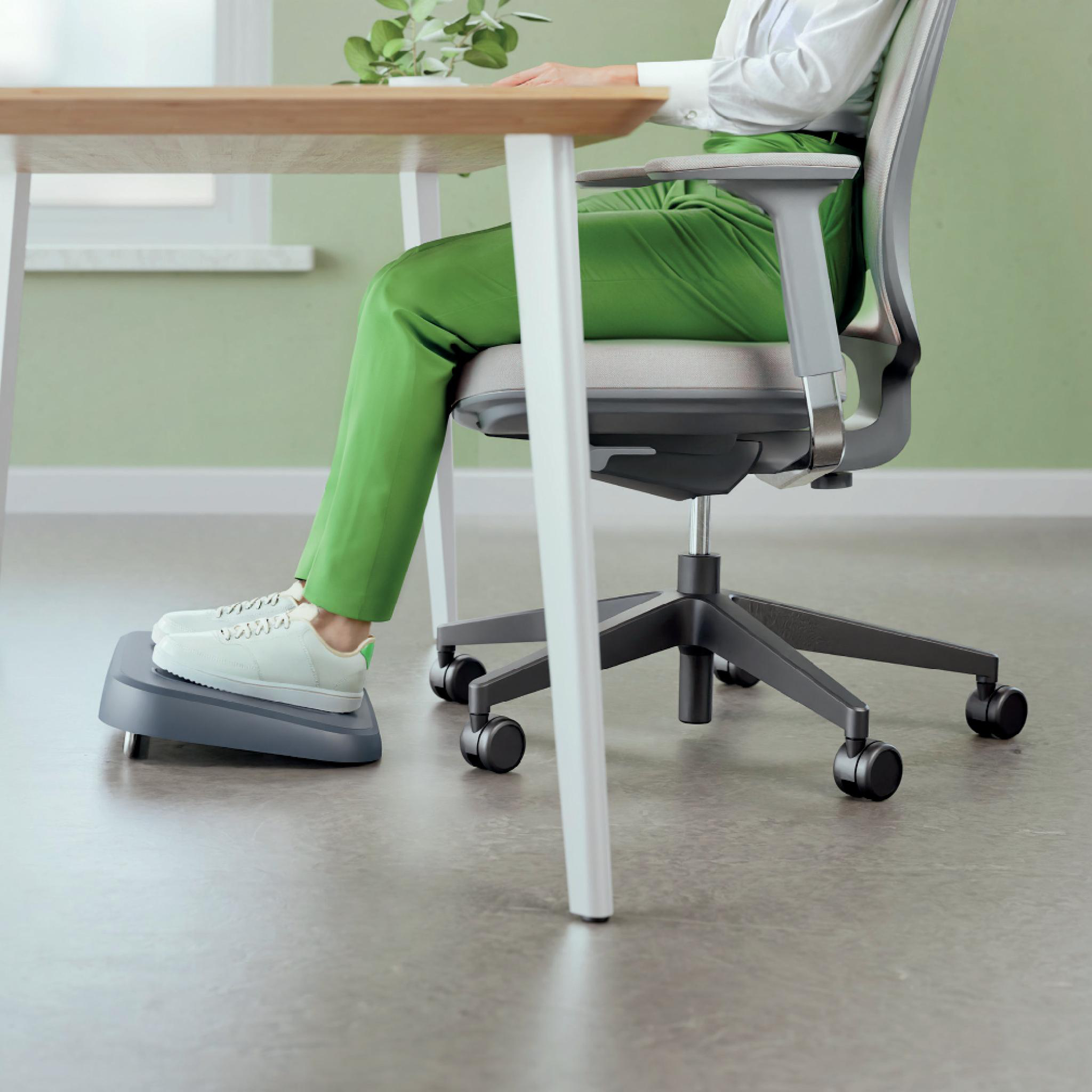 Person in grüner Hose sitzt an einem Schreibtisch mit Fußstütze. Weiße Schuhe liegen auf der Fußstütze. Bürostuhl und Tisch sind im Bild.