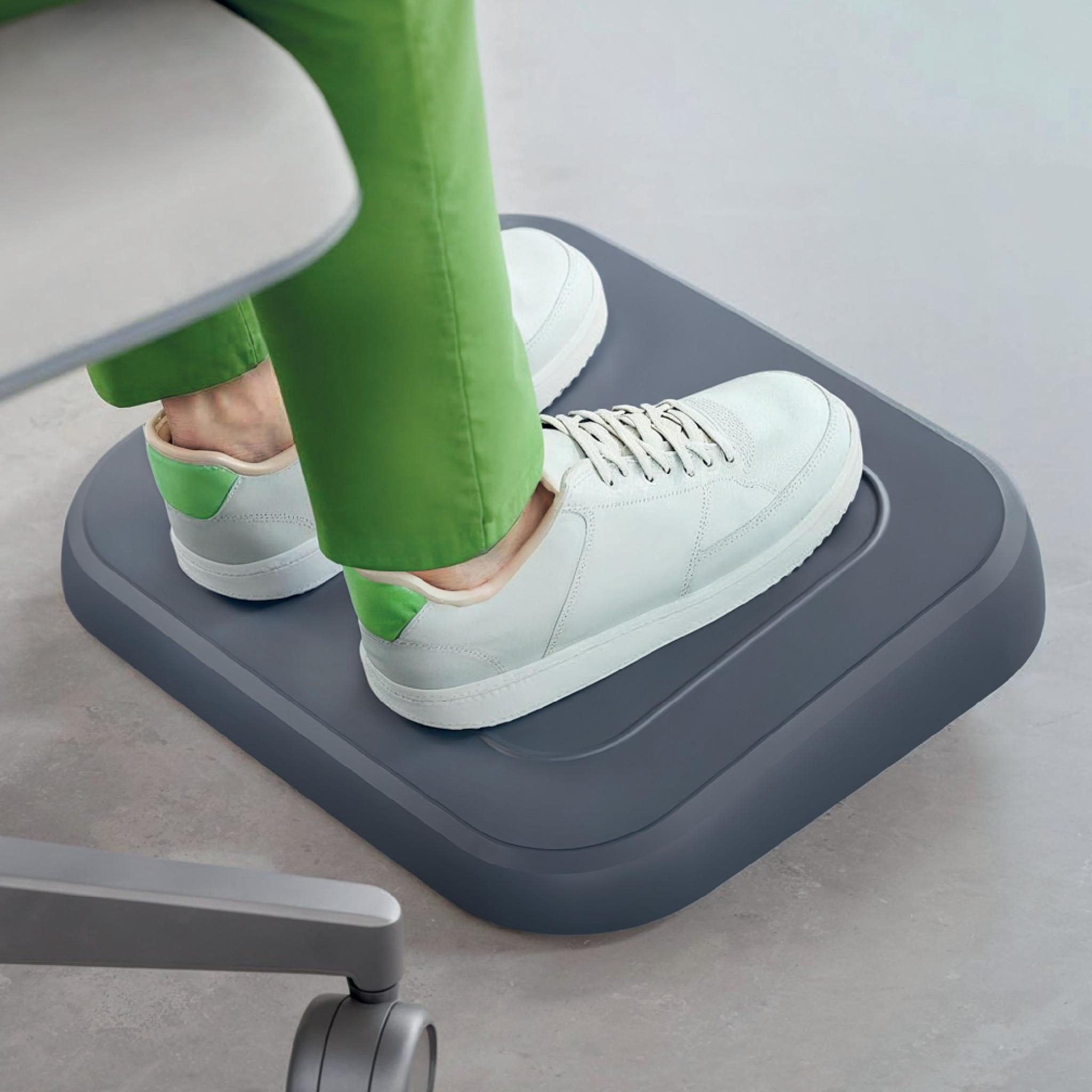 Füße mit weißen Turnschuhen und grüner Hose auf einer grauen Fußstütze. Ein Stuhl ist sichtbar.
