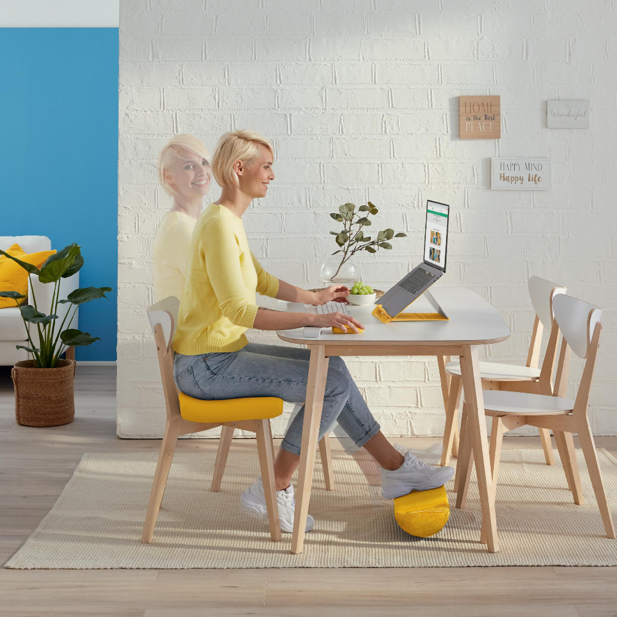 Frau arbeitet am Laptop. Gelber Stuhl und Fußstütze. Weiße Ziegelwand mit Dekoschildern.
