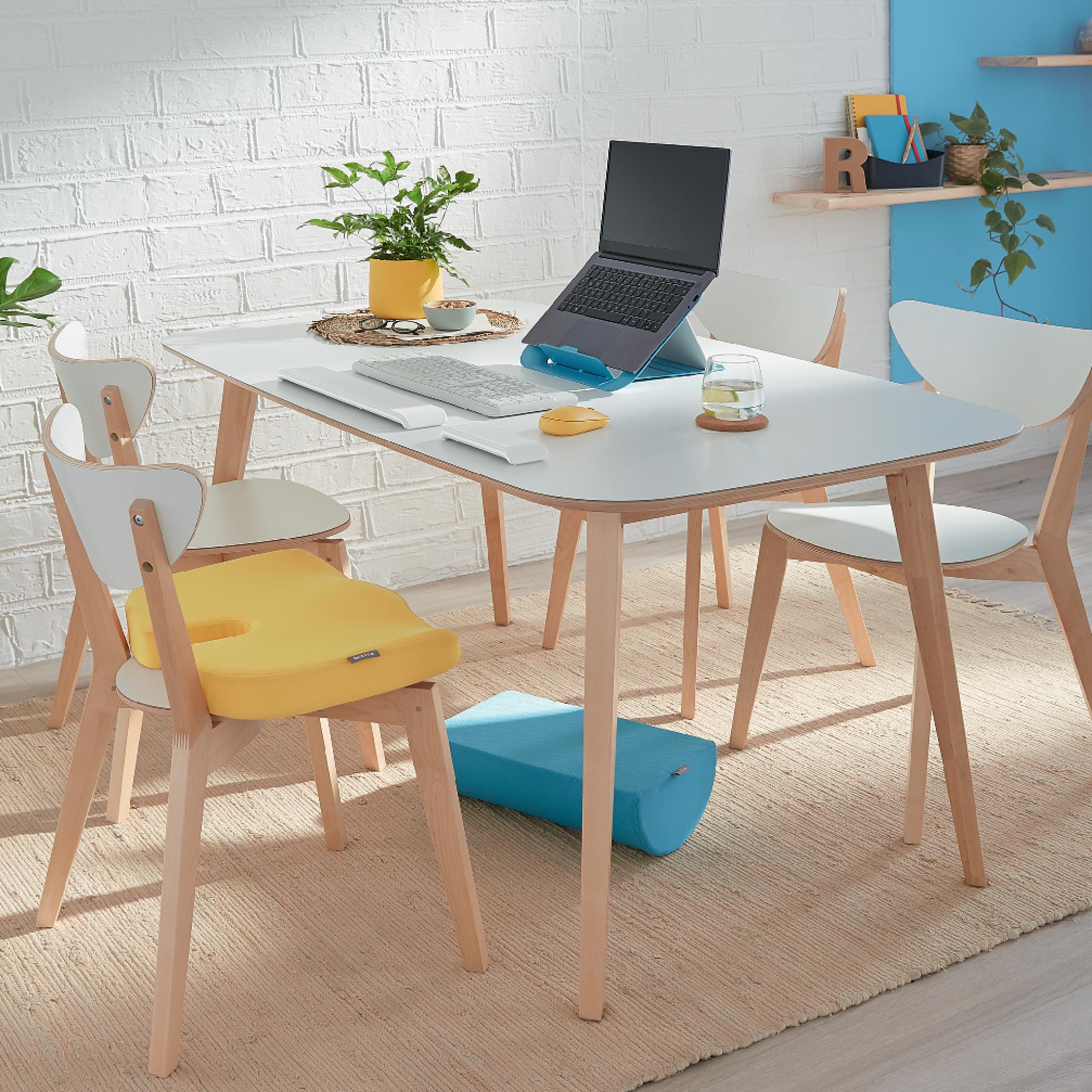 Ein Arbeitsplatz mit weißem Tisch, Laptop und gelbem Stuhlkissen. Enthält Pflanzen, Tastatur und eine blaue Fußstütze.
