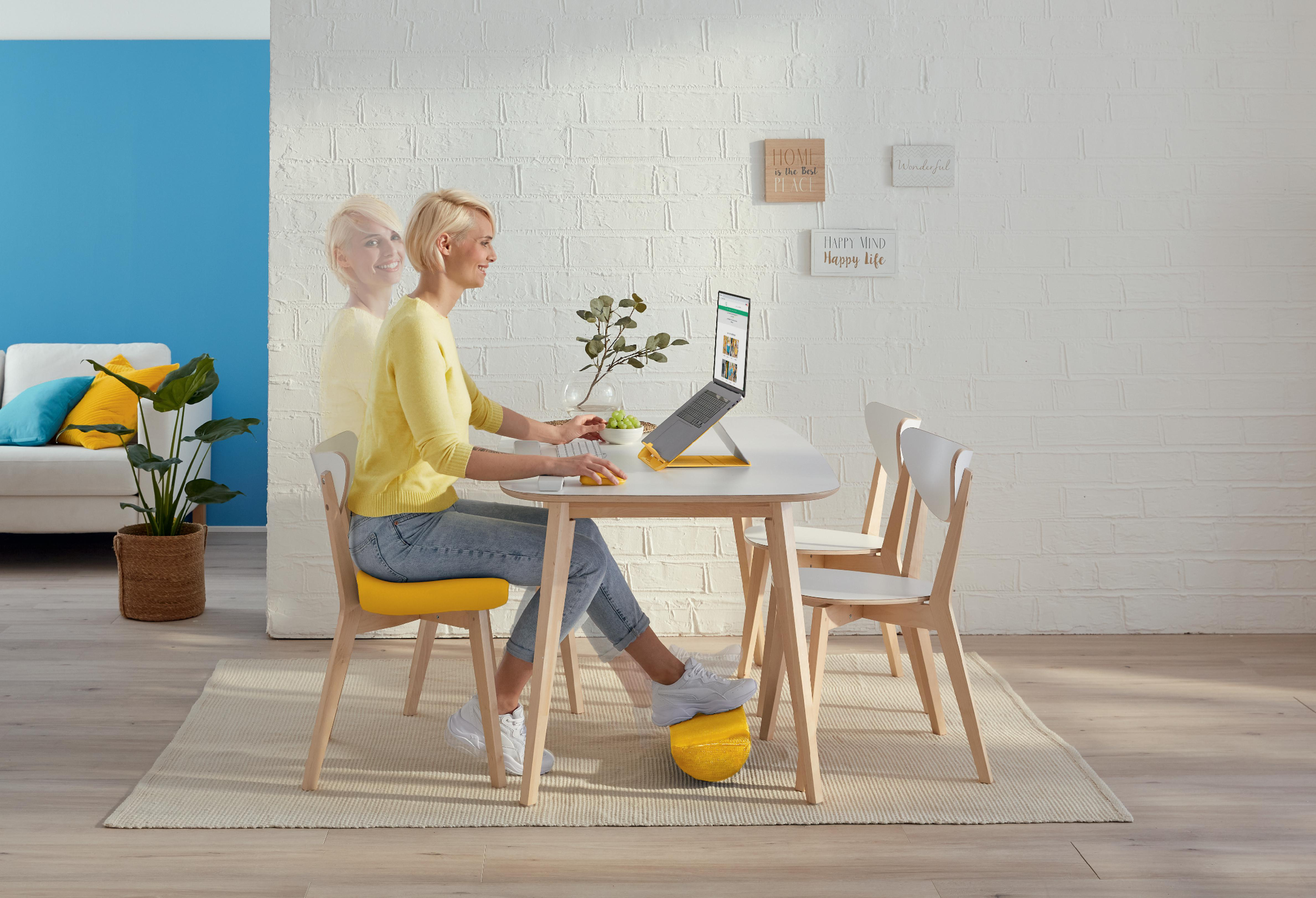 Frau arbeitet am Laptop am Tisch. Weißer Backsteinmauer-Hintergrund. Gelber Stuhl und Fußstütze. Pflanze und Obst in der Nähe.