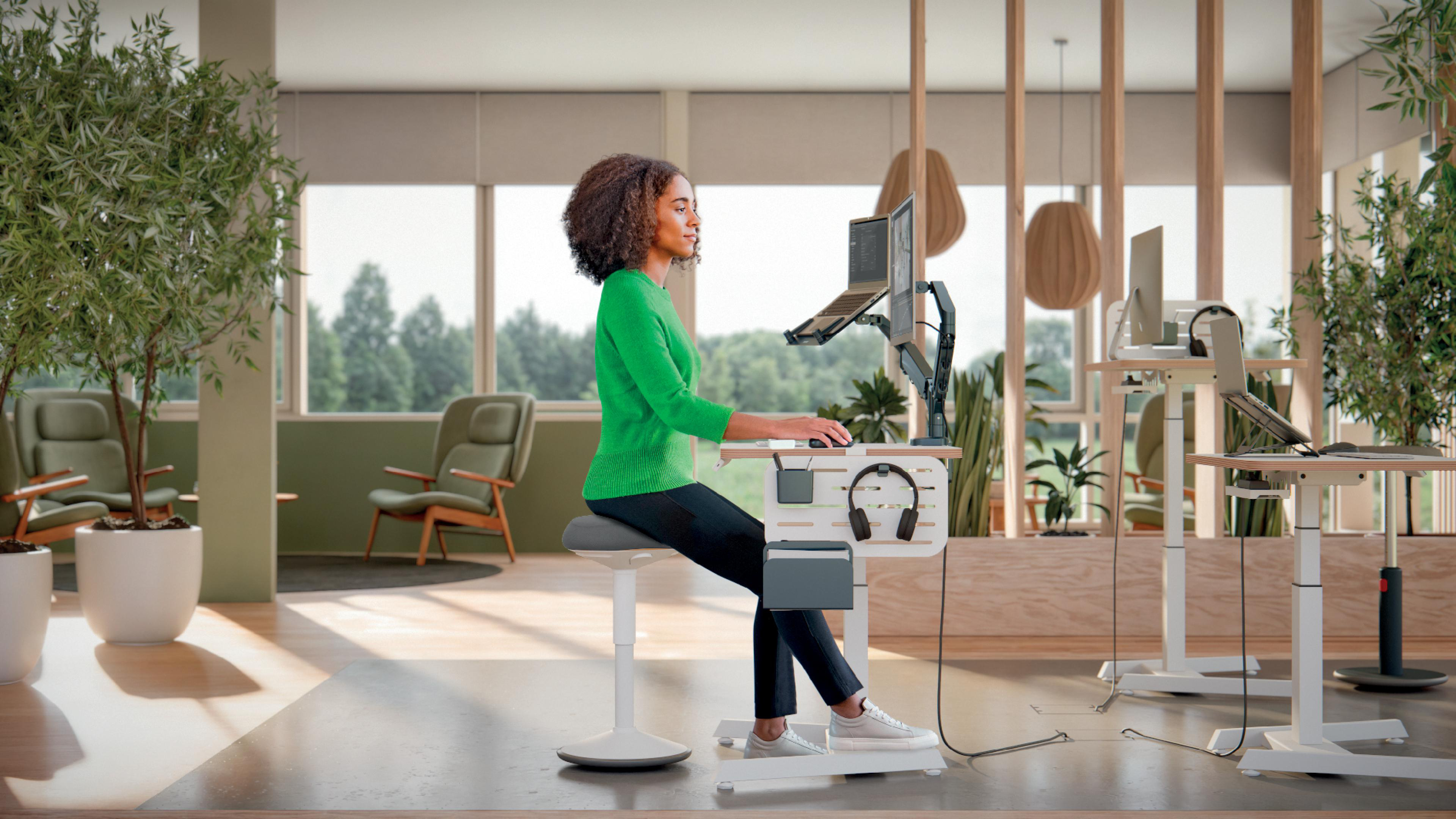 Eine Frau in grünem Pullover arbeitet an einem Schreibtisch. Es gibt Pflanzen, einen Computer und Büromöbel in einem modernen Büro.