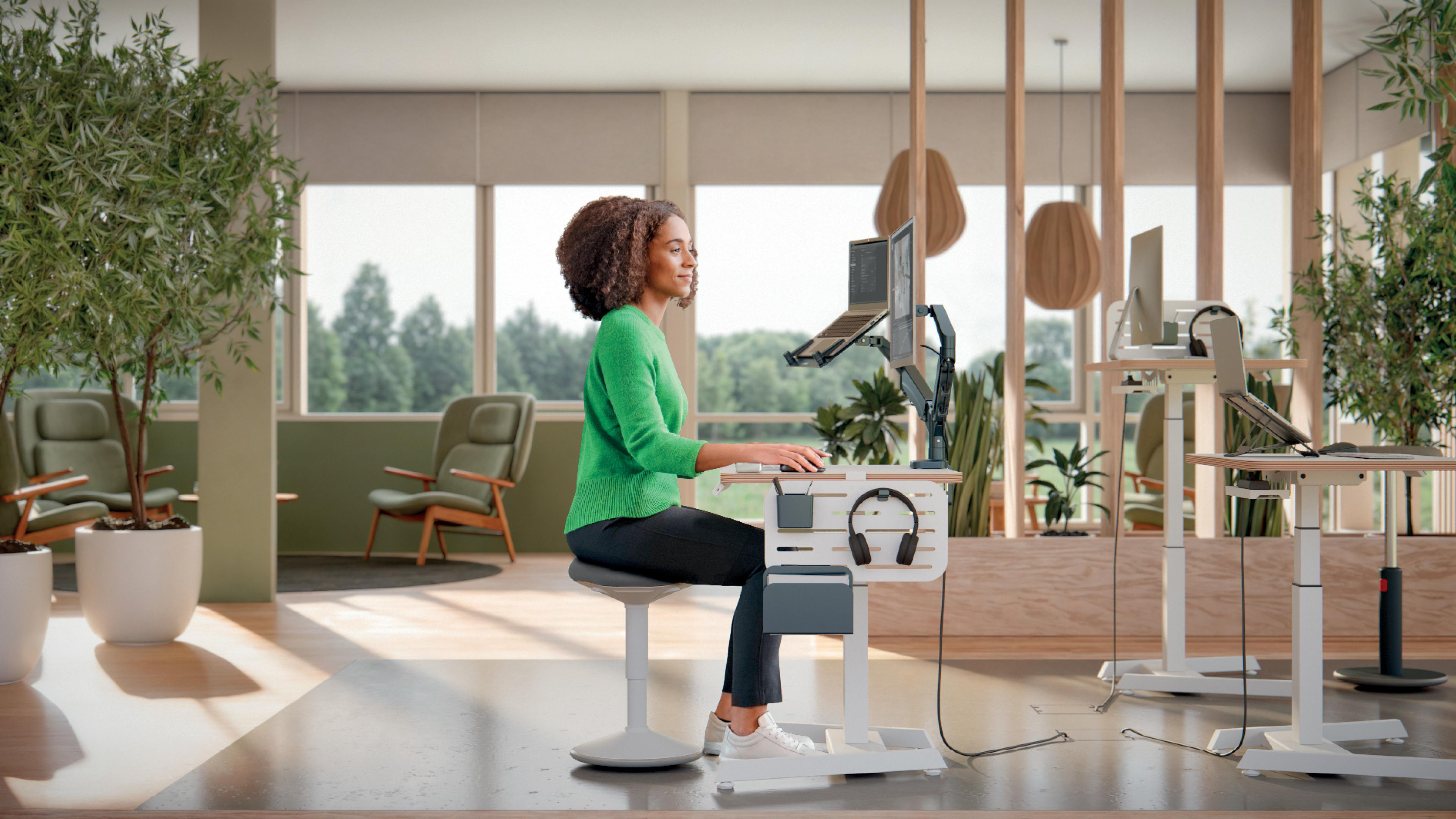 Eine Frau in Grün arbeitet an einem Schreibtisch. Das Büro hat Pflanzen, Monitore und Holzakzente. Es ist ein moderner, heller Arbeitsplatz.