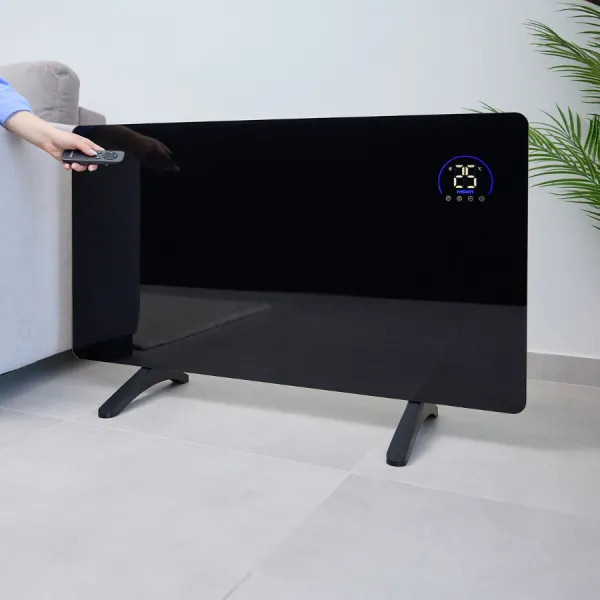 Czarny grzejnik z cyfrowym wyświetlaczem pokazuje 25 stopni. Ręka trzyma pilota. Po prawej roślina.