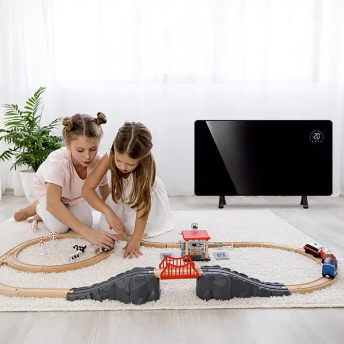 Dwie dziewczynki bawią się pociągiem zabawkowym na dywanie. Z tyłu stoi czarny grzejnik. Roślina po lewej.