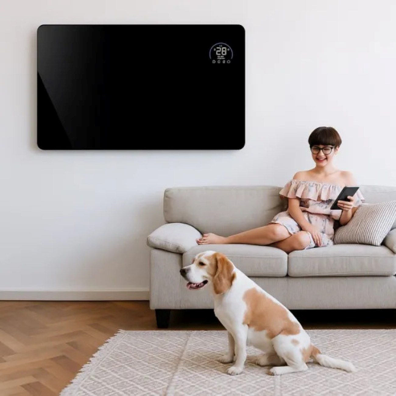 Kobieta siedzi na sofie, trzymając tablet, z psem przed sobą. Czarny grzejnik panelowy jest zamontowany na ścianie.