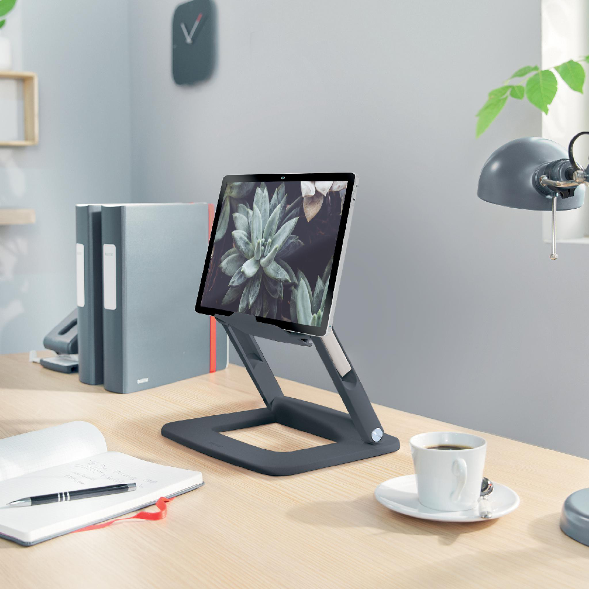Tablet auf einem Ständer, Kaffeetasse, Notizbuch und Lampe auf einem Holztisch. Graue Wand und Uhr im Hintergrund.