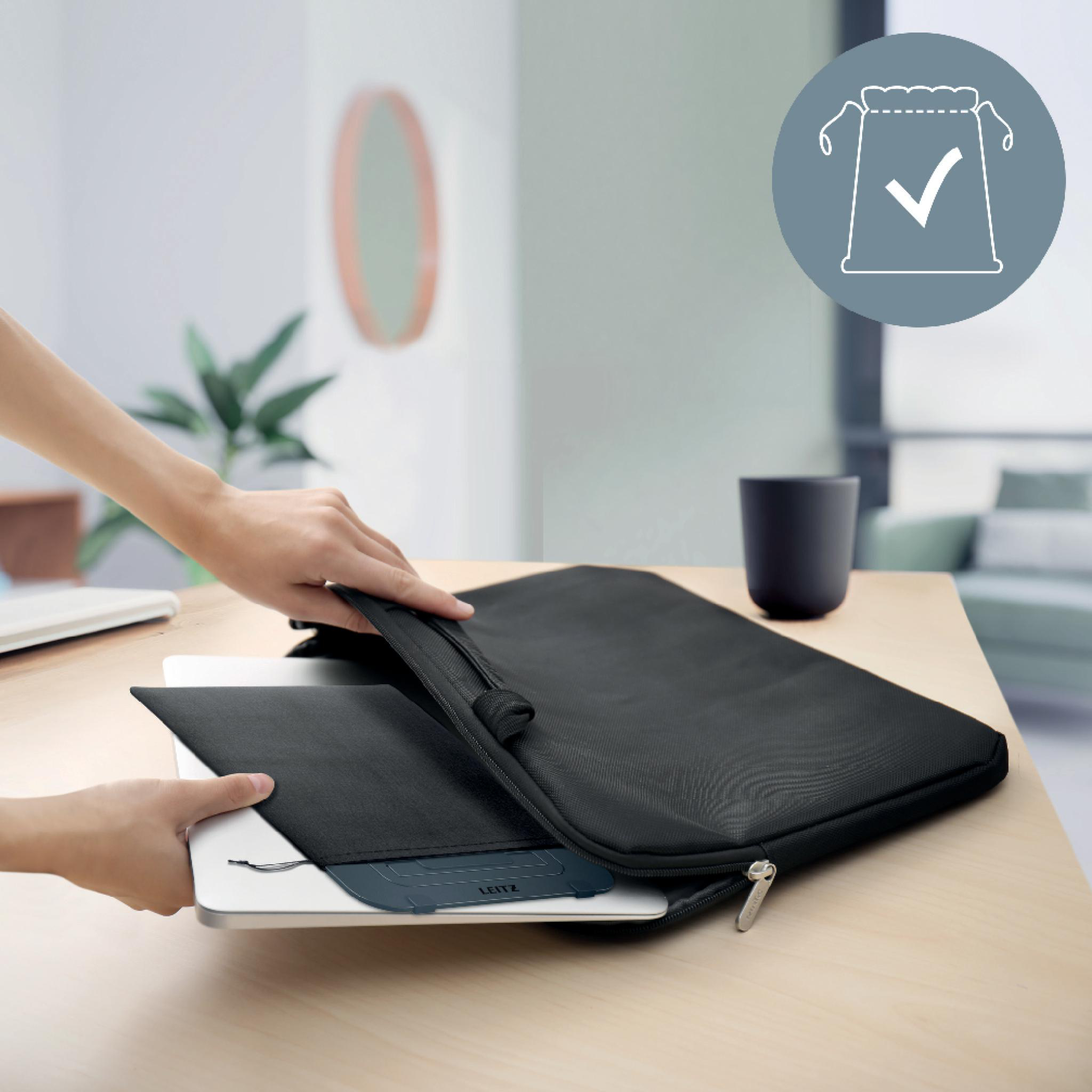 Eine Person nimmt einen Laptop aus einer schwarzen Laptoptasche auf einem Holztisch. Eine Tasse und eine Wanddekoration sind sichtbar.